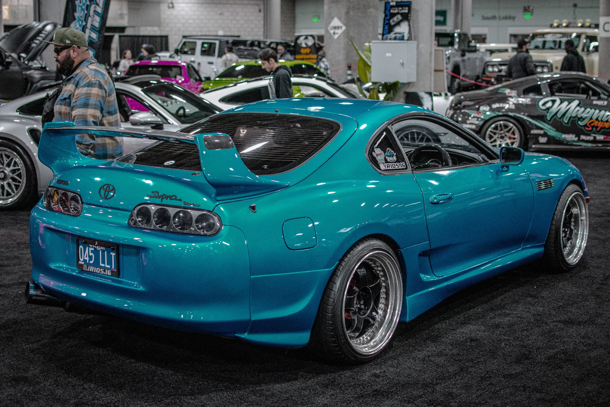 A bright blue Toyota Supra sports car with a large rear spoiler on display at an indoor car show, surrounded by other modified vehicles and people viewing.