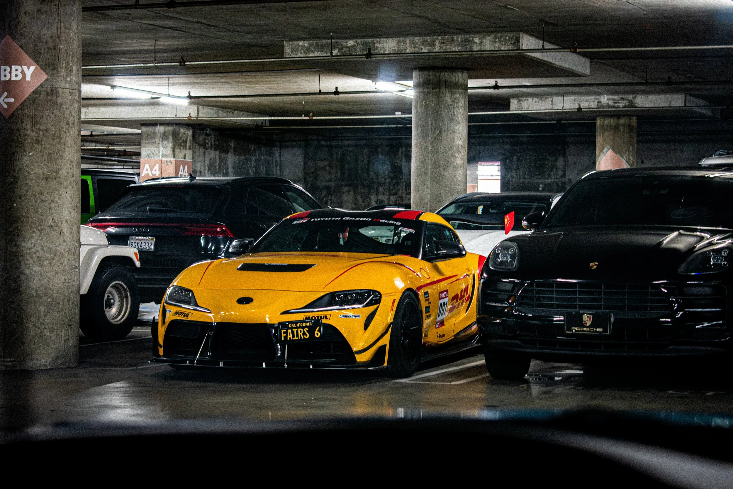 Yellow race car with racing decals parked in an underground parking garage surrounded by black and white luxury cars.
