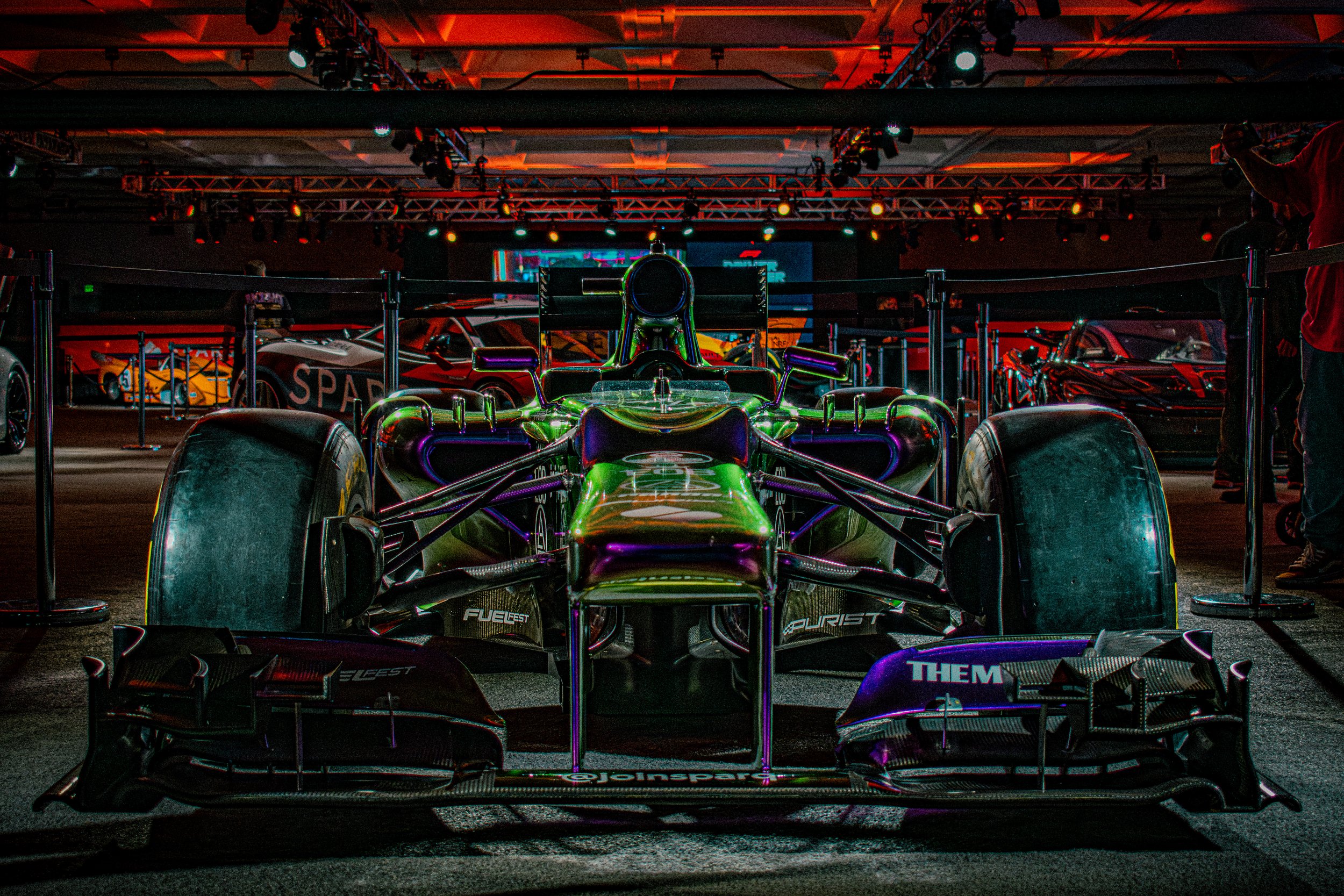 A race car displayed at an exhibition with several other race cars in the background under colorful lighting.