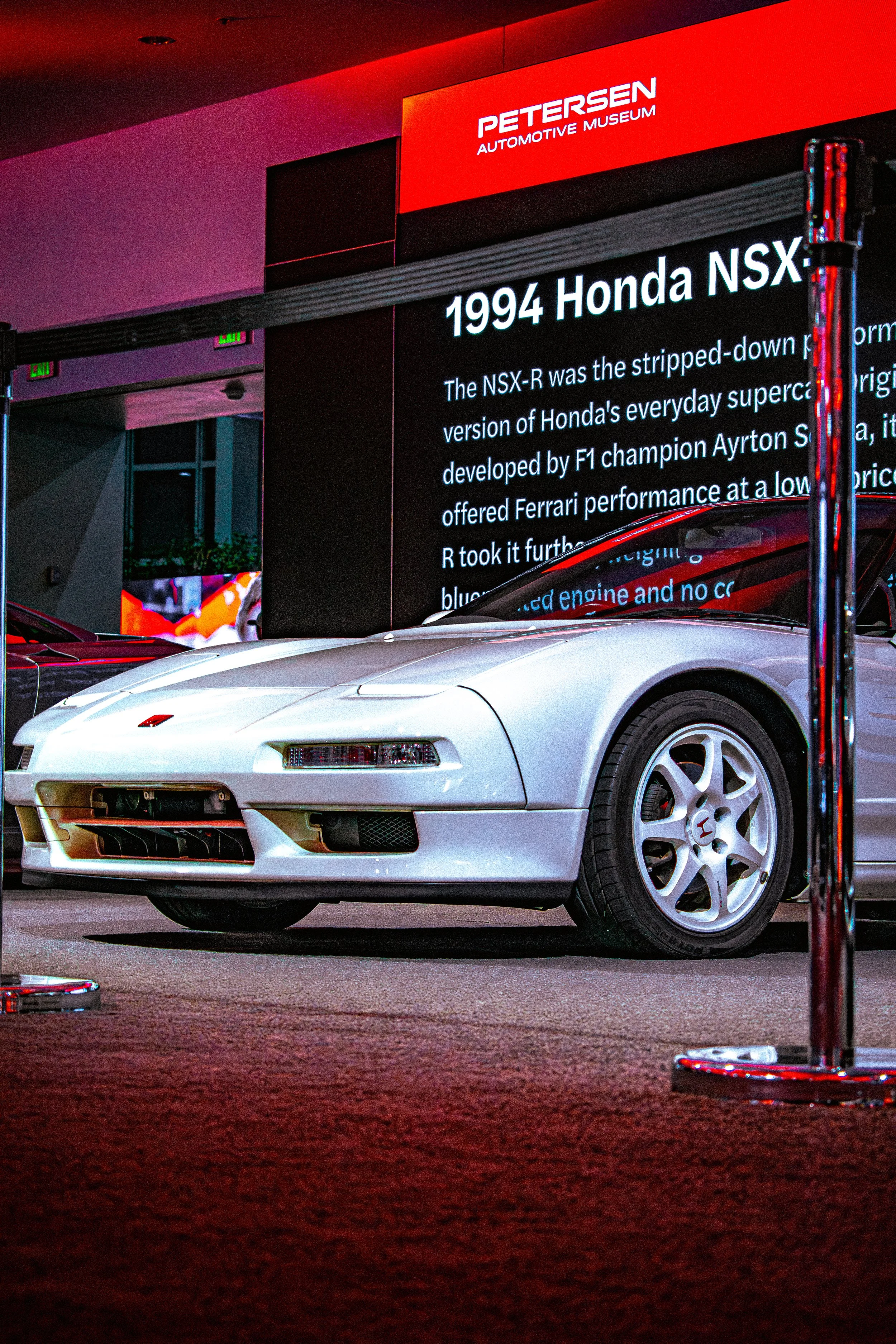 A white 1994 Honda NSX sports car on display at Petersen Automotive Museum, with an information screen behind it highlighting details about the car.