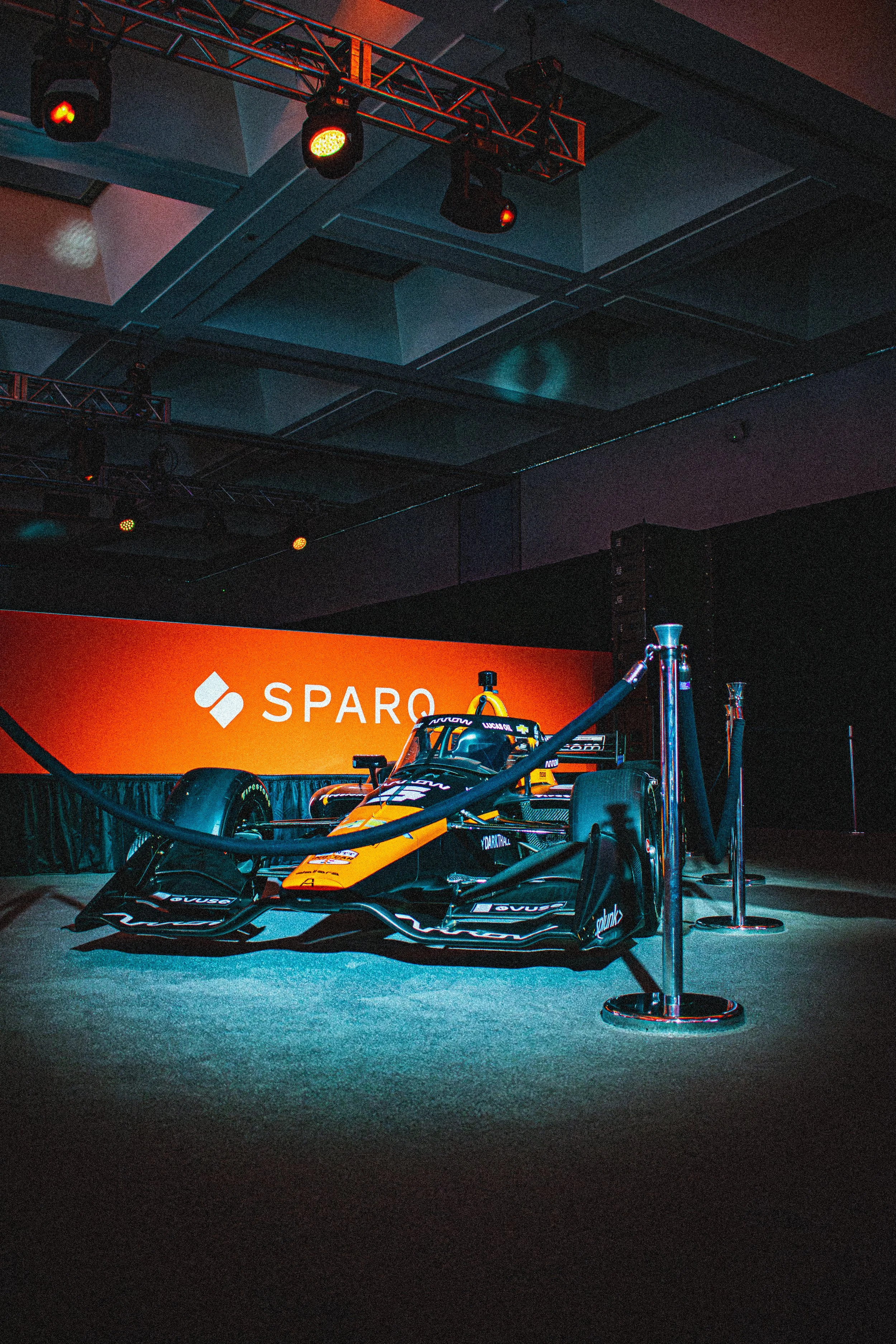 A race car displayed on a stage with a bright orange banner behind it, featuring the Sparco logo and text, under dark lighting with spotlights above.