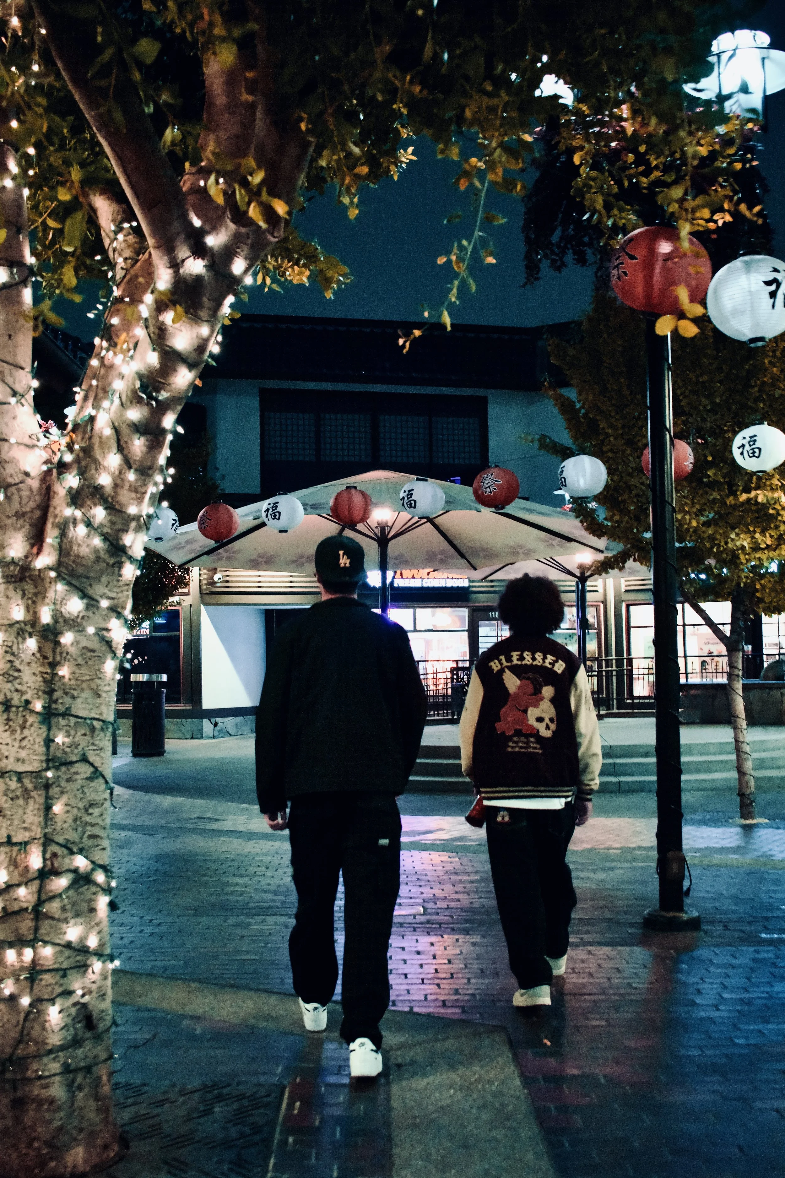 Two young men walk under decorative red and white lanterns in an outdoor area at night, illuminated by string lights and streetlights.