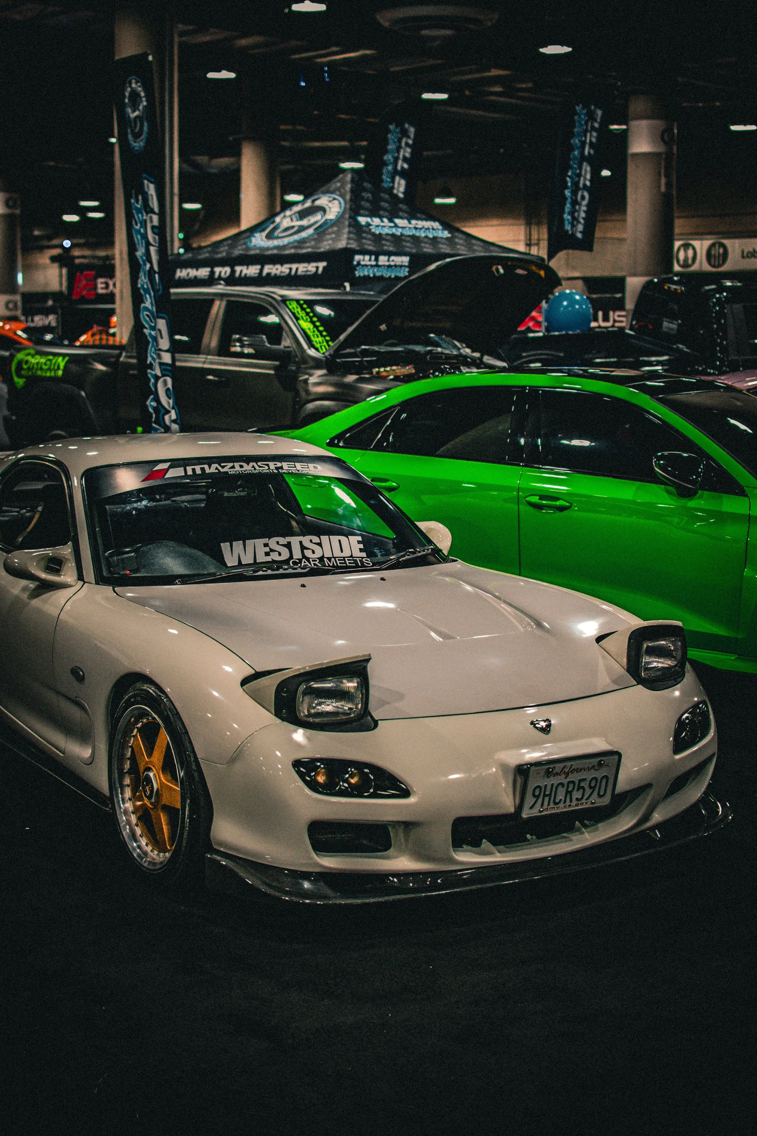 A white MazdaSpeed sports car with yellow wheels and a front lip, displayed at an indoor car show alongside a green car and other vehicles, with banners and tents in the background.