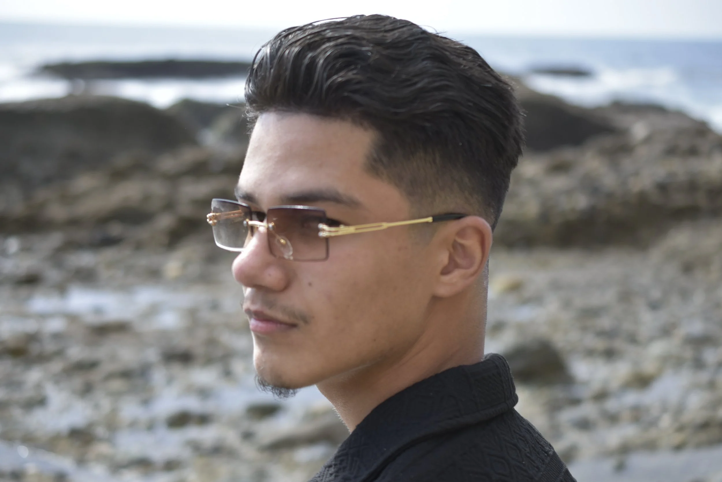 A young man with short, styled black hair and light skin wearing sunglasses and a black shirt at a rocky beach.