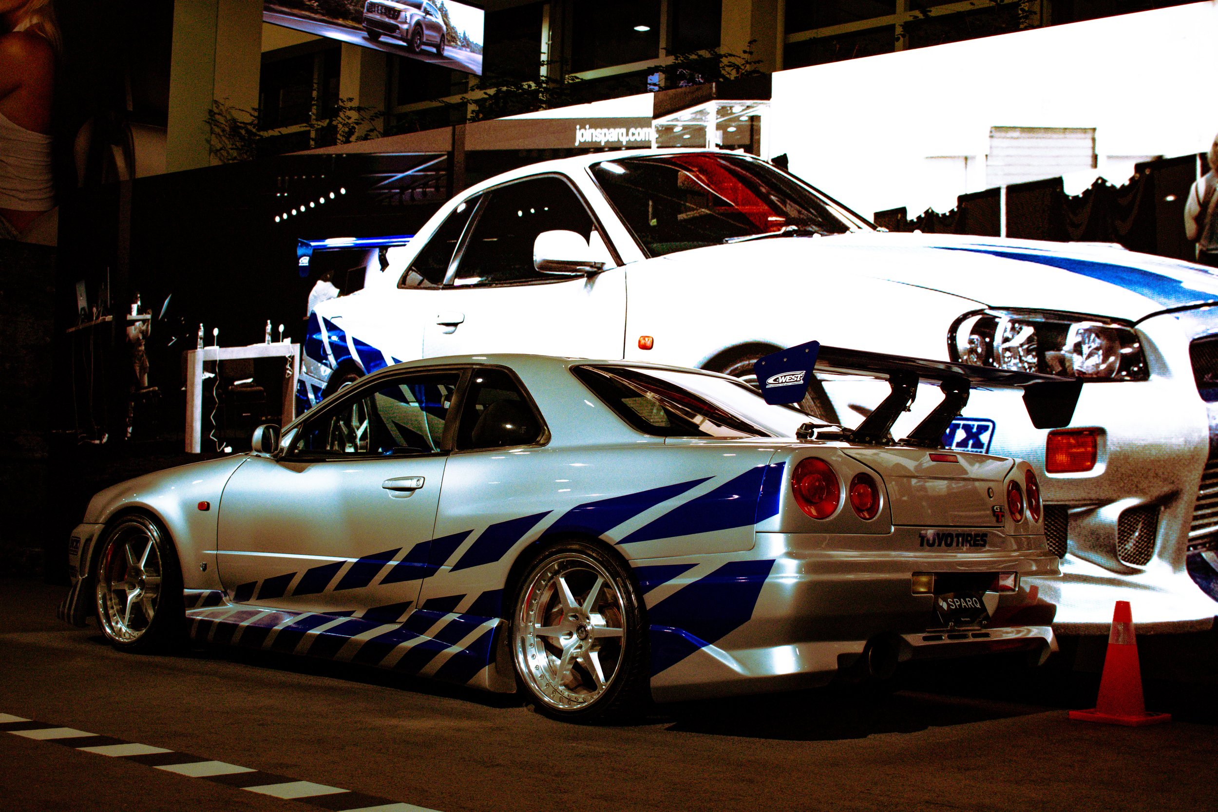 A silver and blue race car with a large rear wing and aftermarket wheels displays at an indoor car exhibition, with another white car in the background.