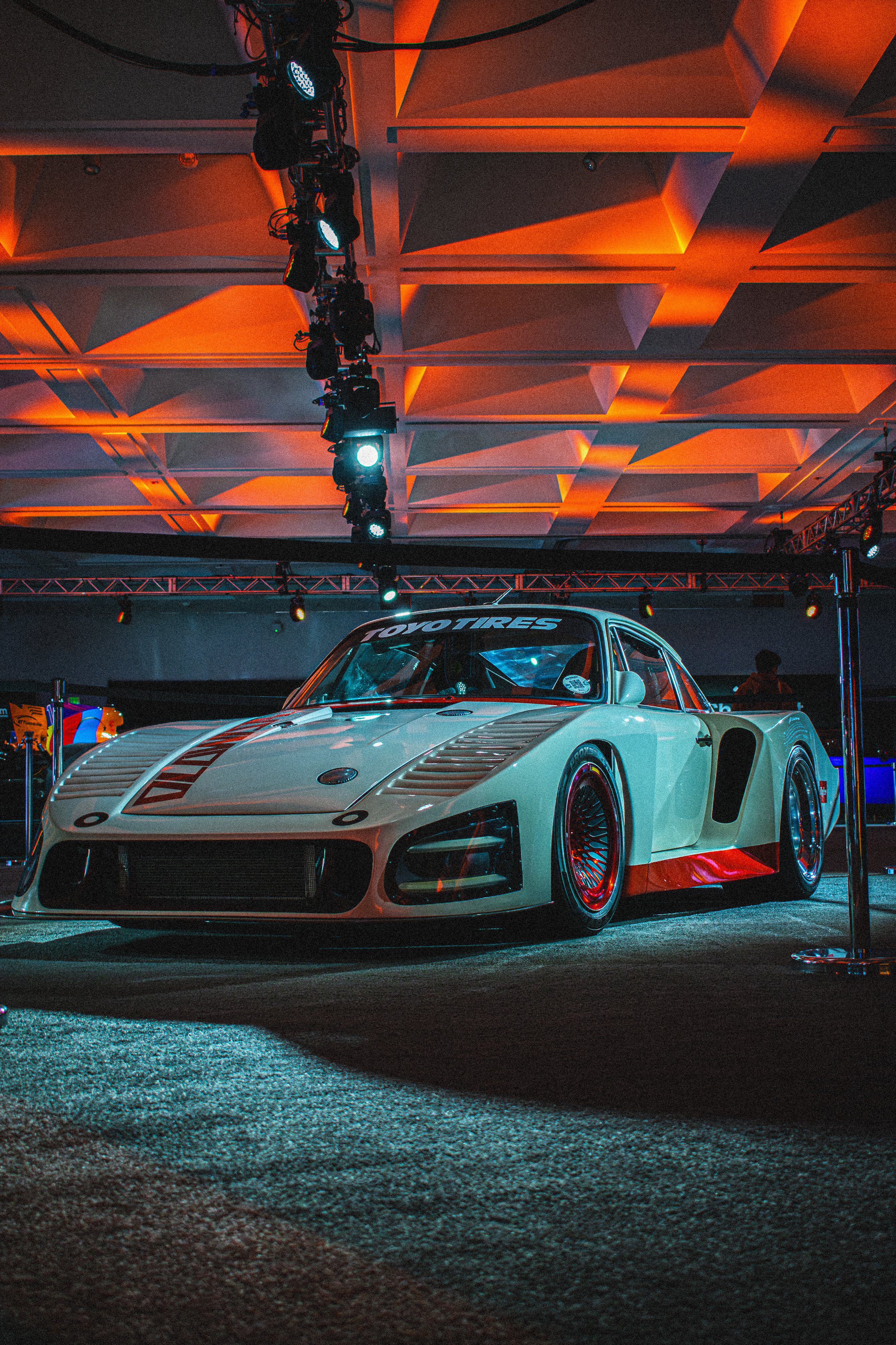 A vintage race car displayed indoors under colorful stage lighting, featuring a sleek design, prominent air vents, and red accents, with a 