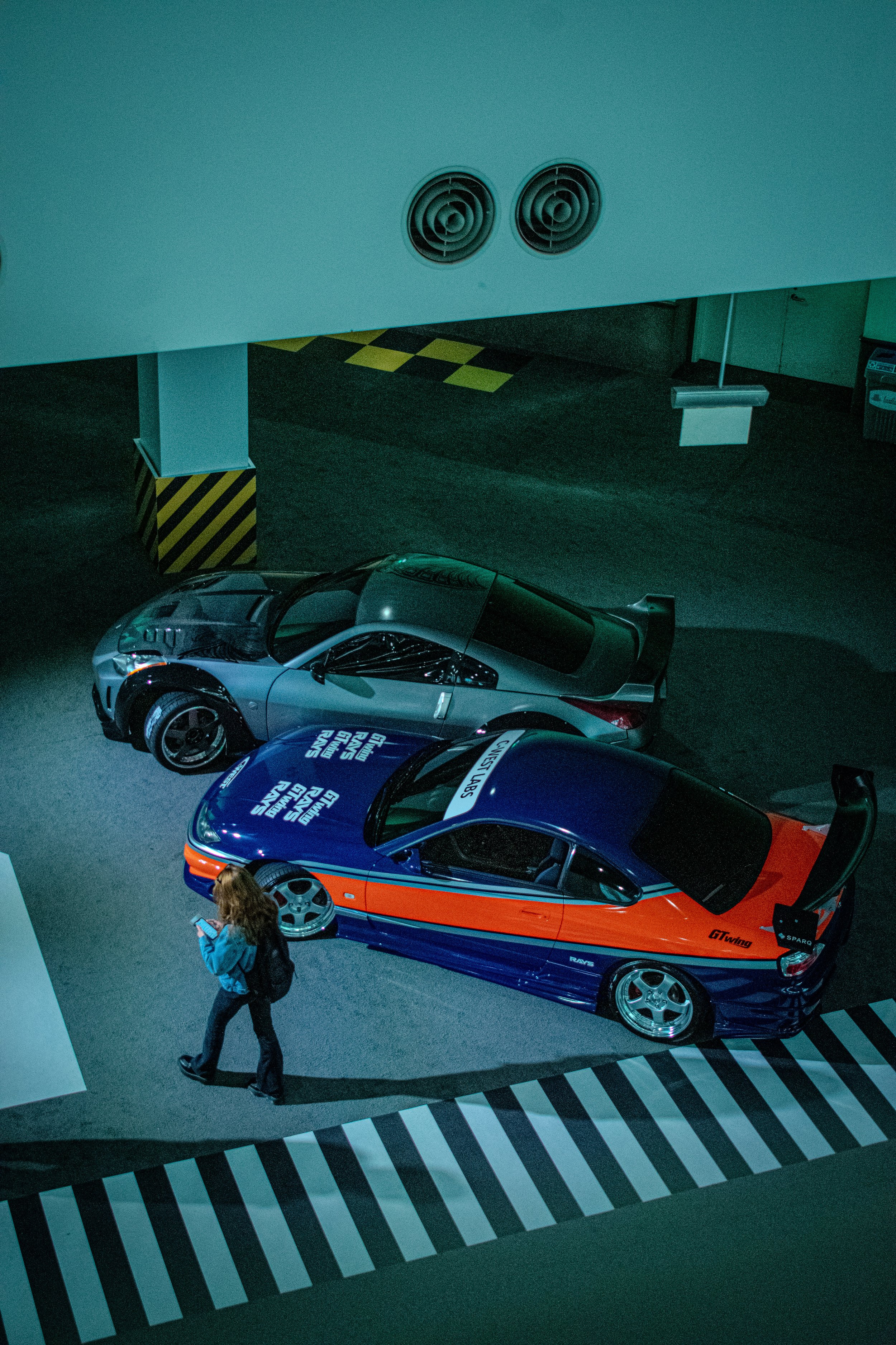 Two race cars, one silver and one blue with orange accents, parked under an overpass next to a pedestrian walking by with a phone in hand.