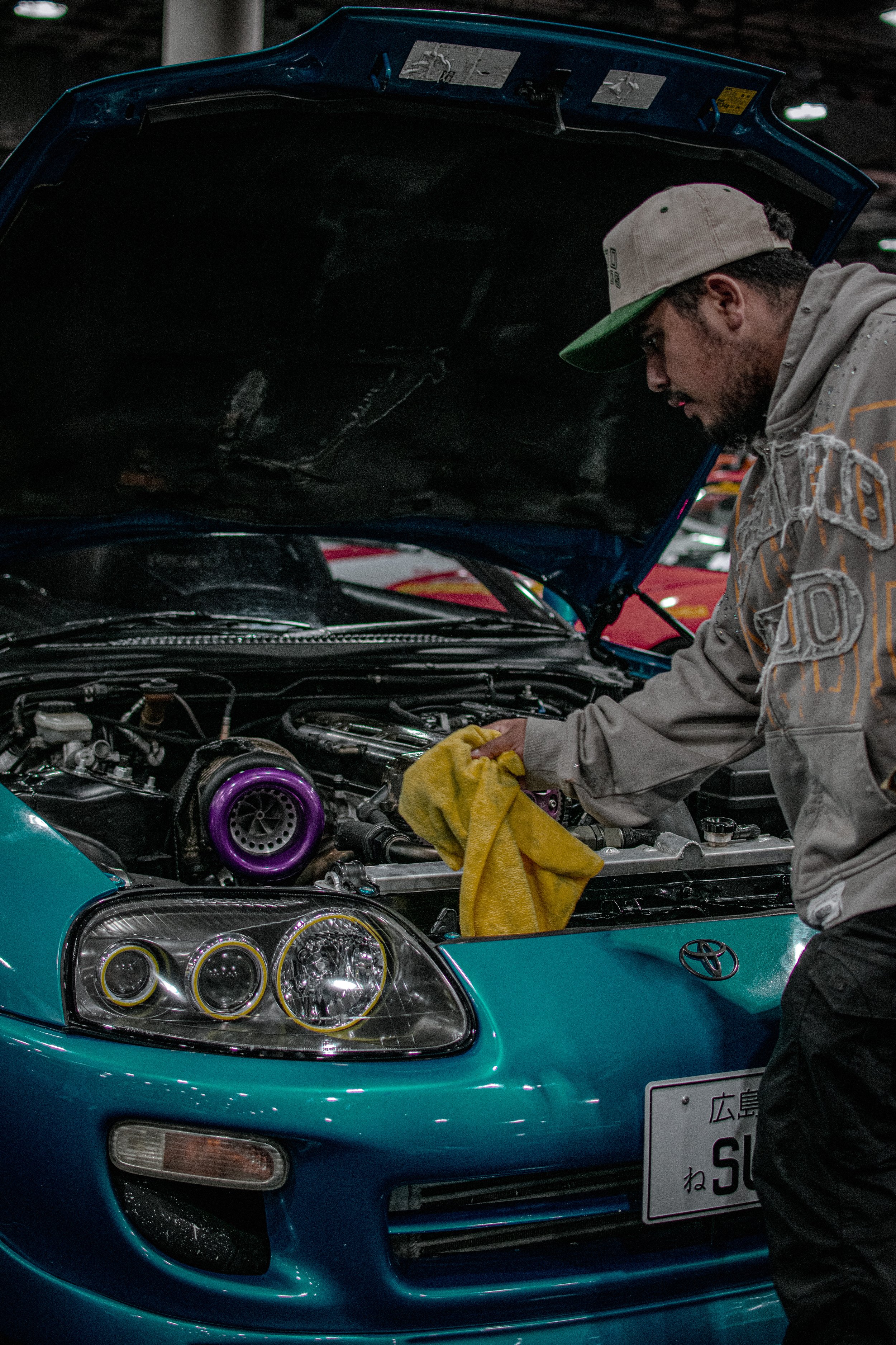 A man wearing a gray hoodie, baseball cap, and black pants is working on the engine of a teal sports car with the hood open at an indoor car show or garage.