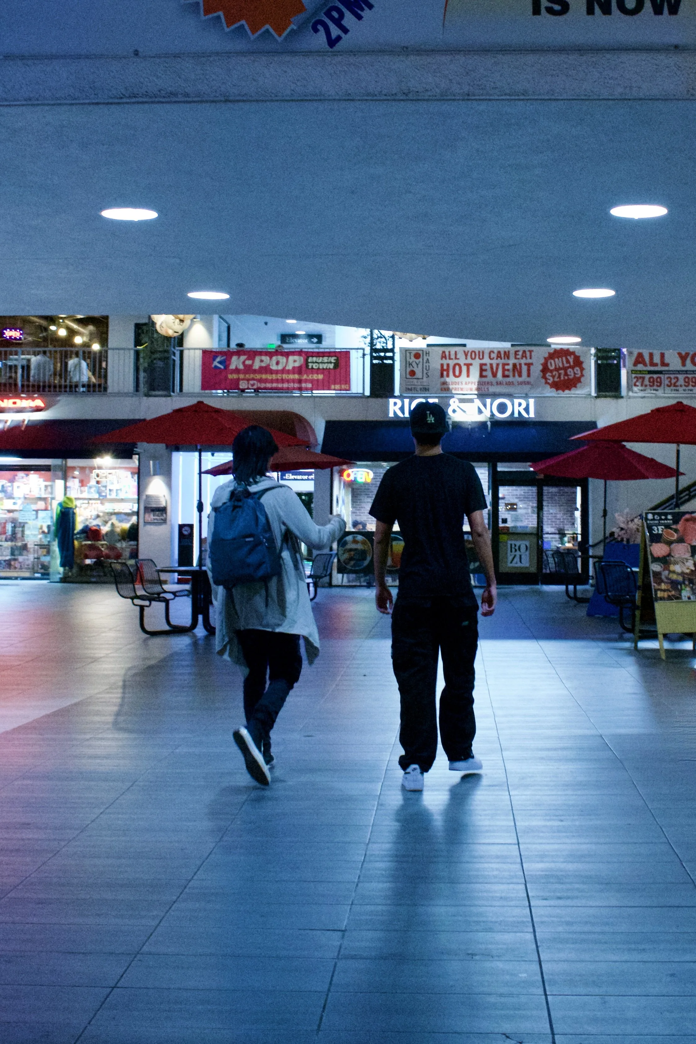 Two people walking through a shopping mall at night, illuminated by blue lighting, with storefronts and signs in the background, some of which promote food and entertainment options.