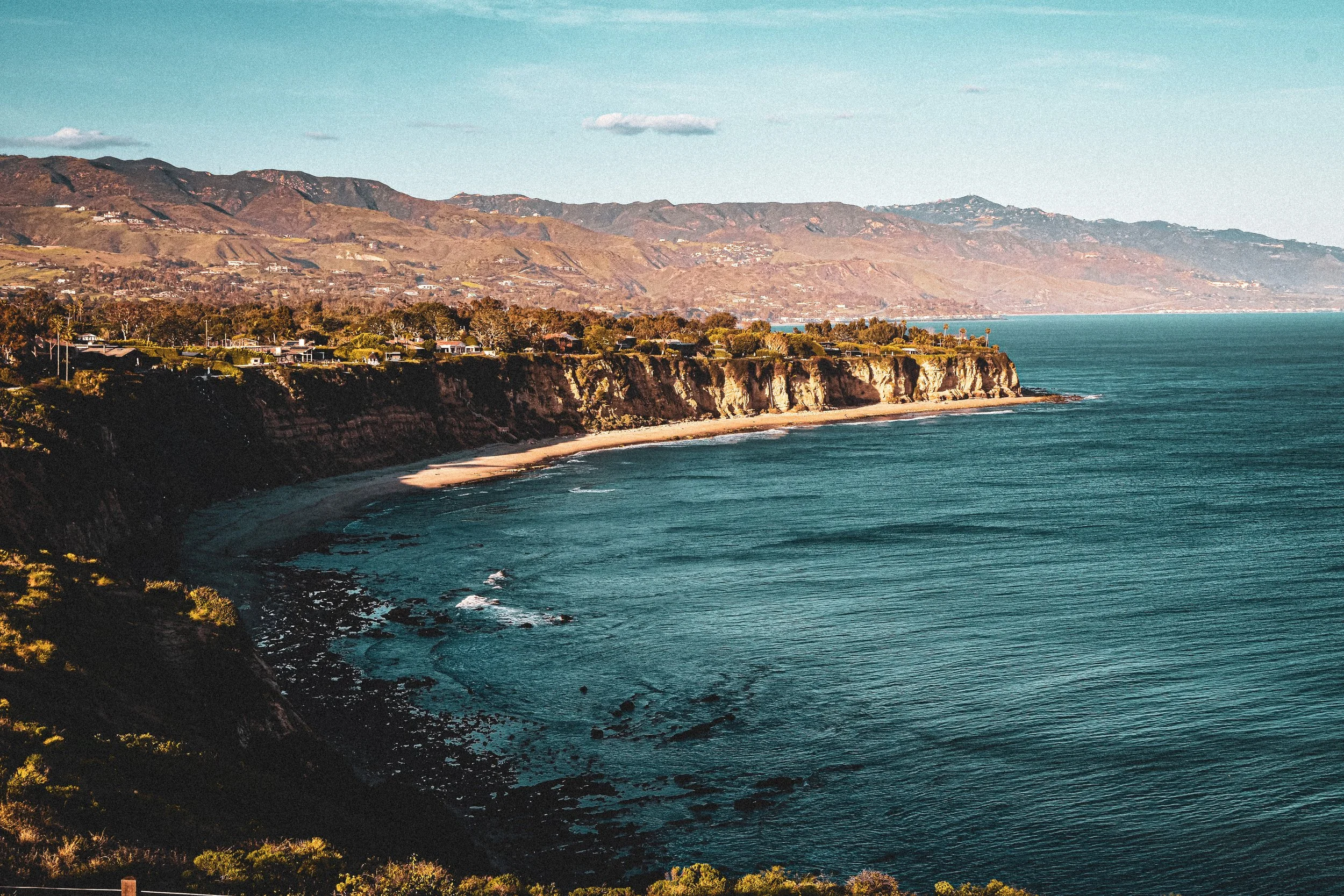 Coastal cliffs overlooking a beach and the ocean with hills in the background and a partly cloudy sky.