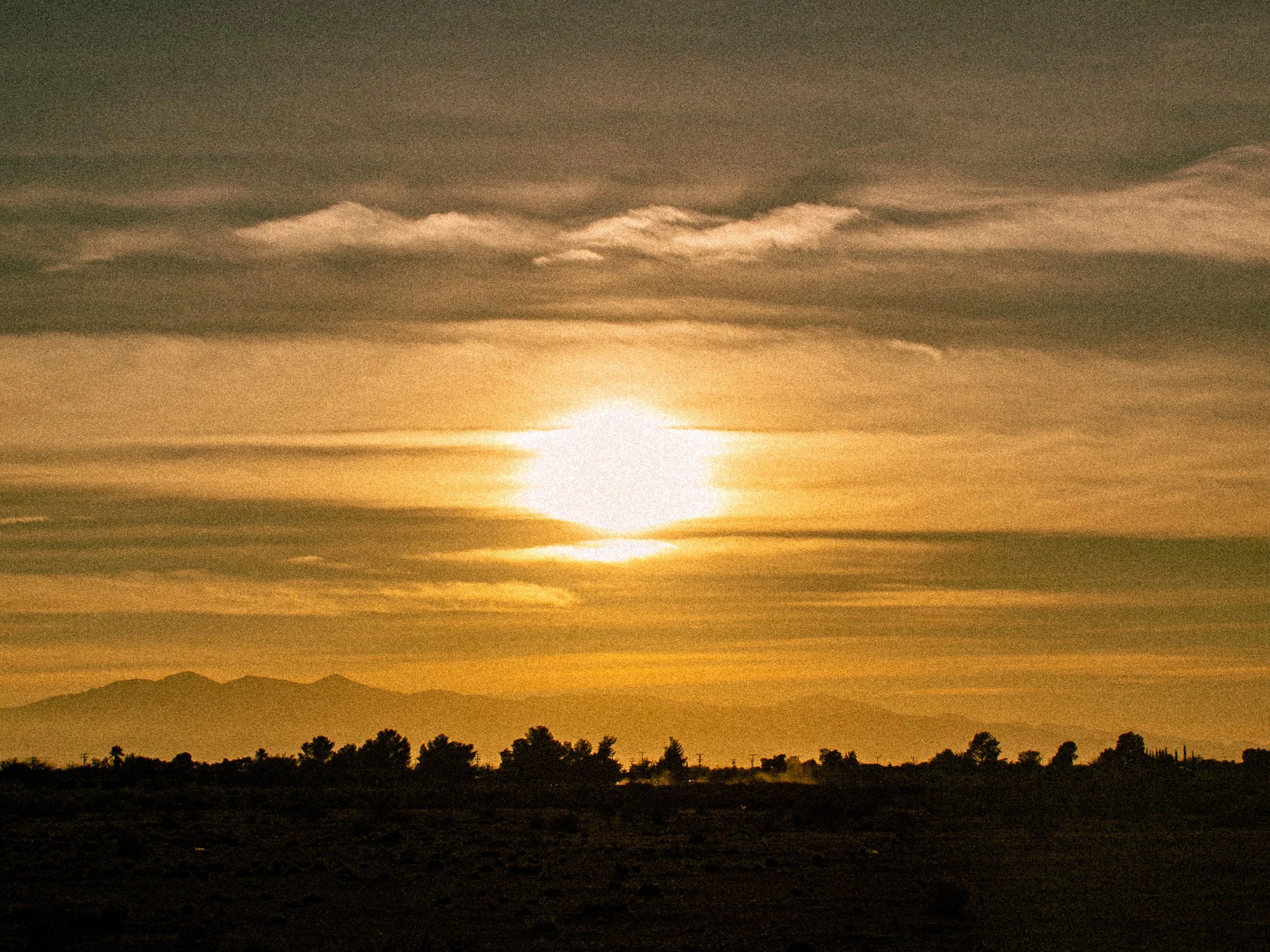 Sunset over a desert landscape with mountains in the background and scattered trees in the foreground.