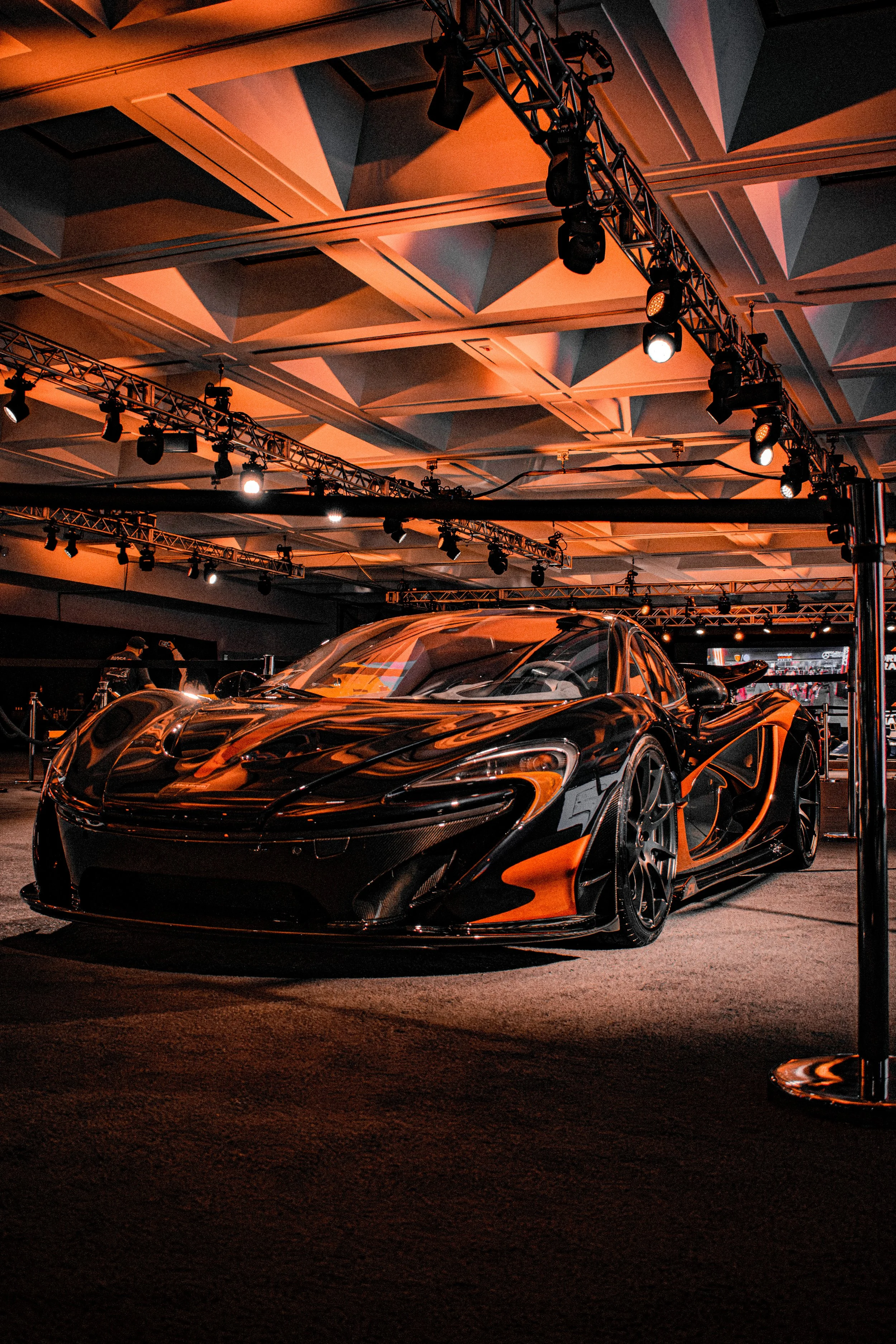 A sleek black sports car on display at an indoor auto show, under orange and black lighting with stage lights and trusses overhead.