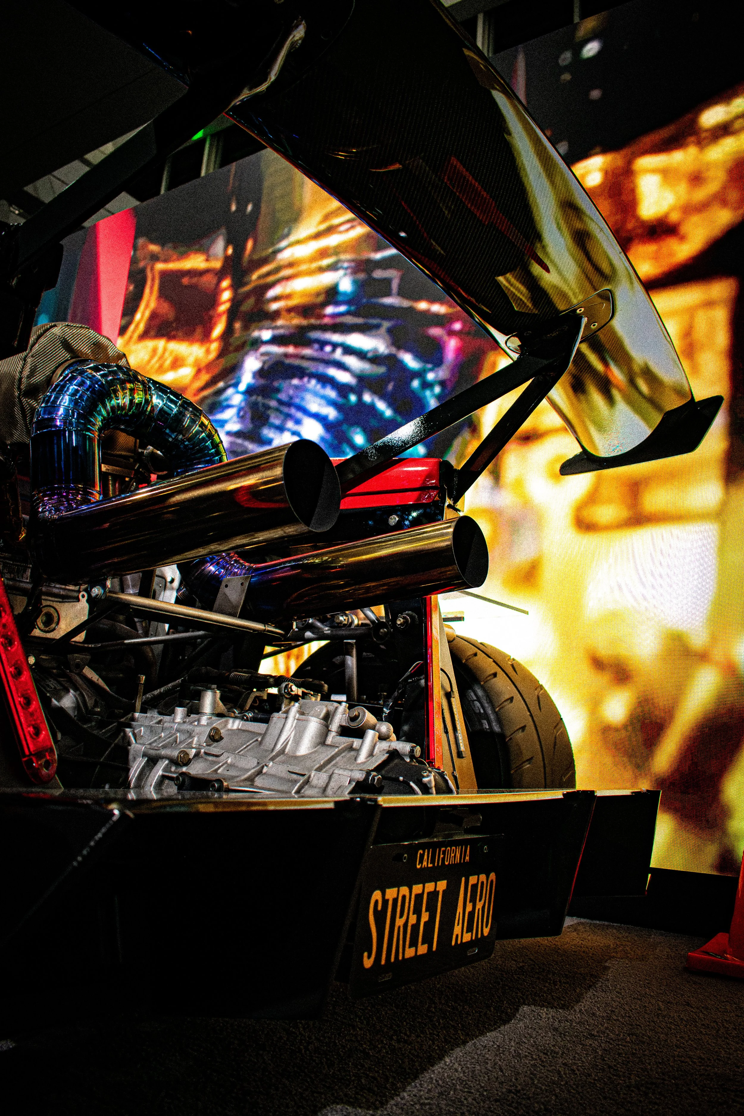 Close-up of a race car with an exposed engine, a large rear wing, and a California license plate reading 'STREET AERO,' set against a vibrant, fiery background on a large screen.