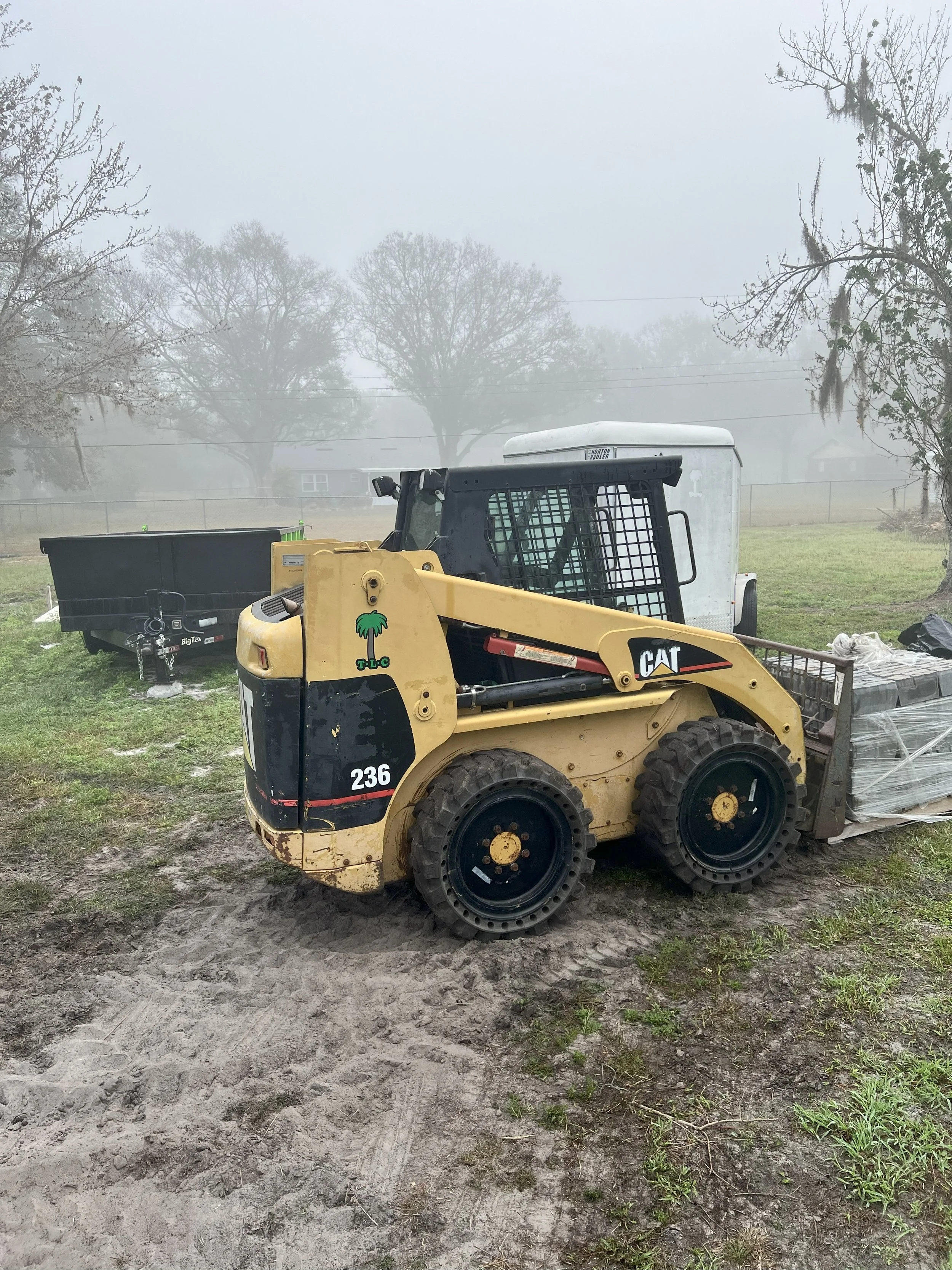 A small yellow CAT skid steer loader sitting on muddy ground with a trailer and open fields in the background, shrouded in fog.