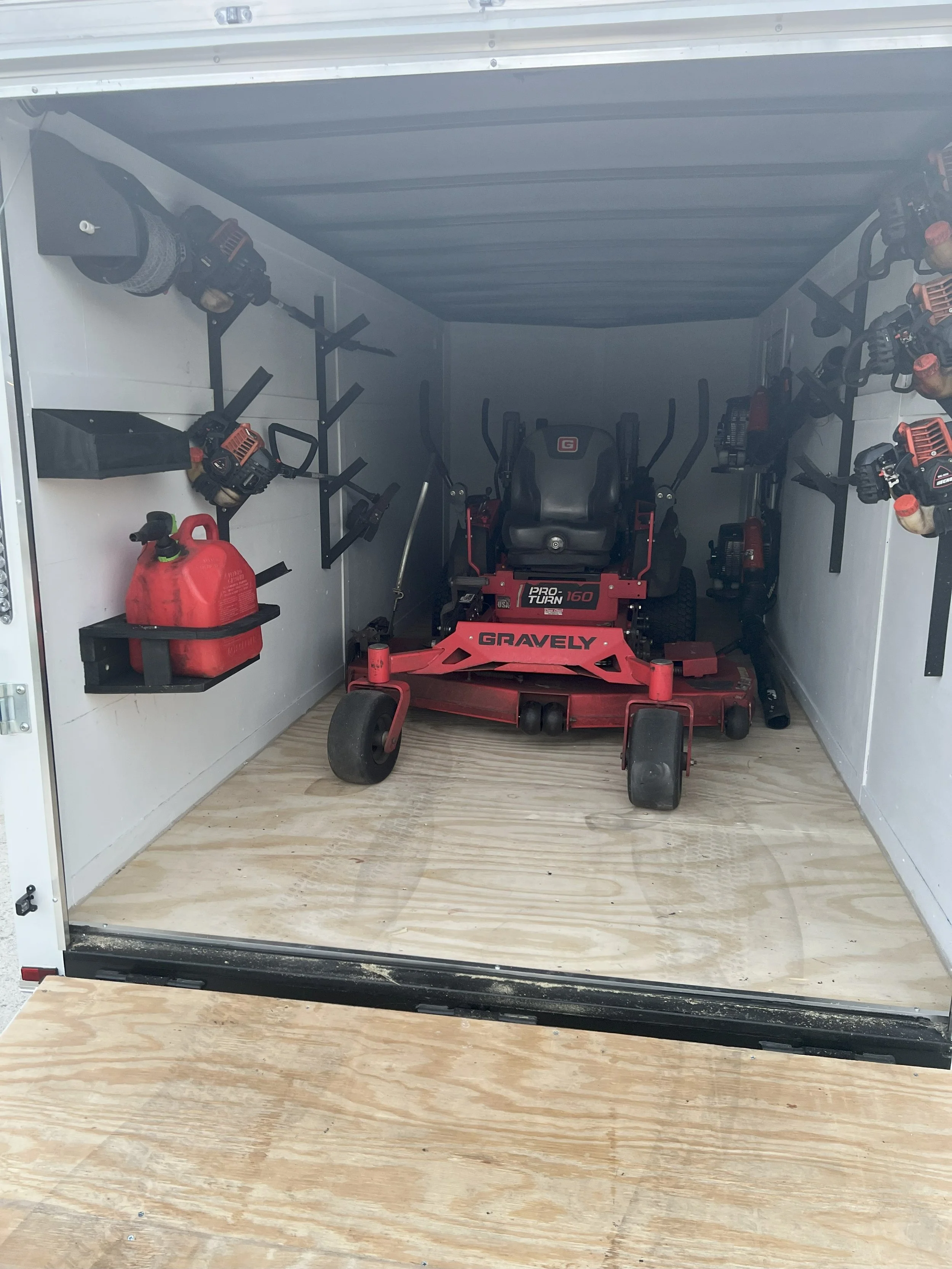 Interior of a mobile tool and equipment storage trailer with a riding mower, power tools on shelves, and a red fuel container.