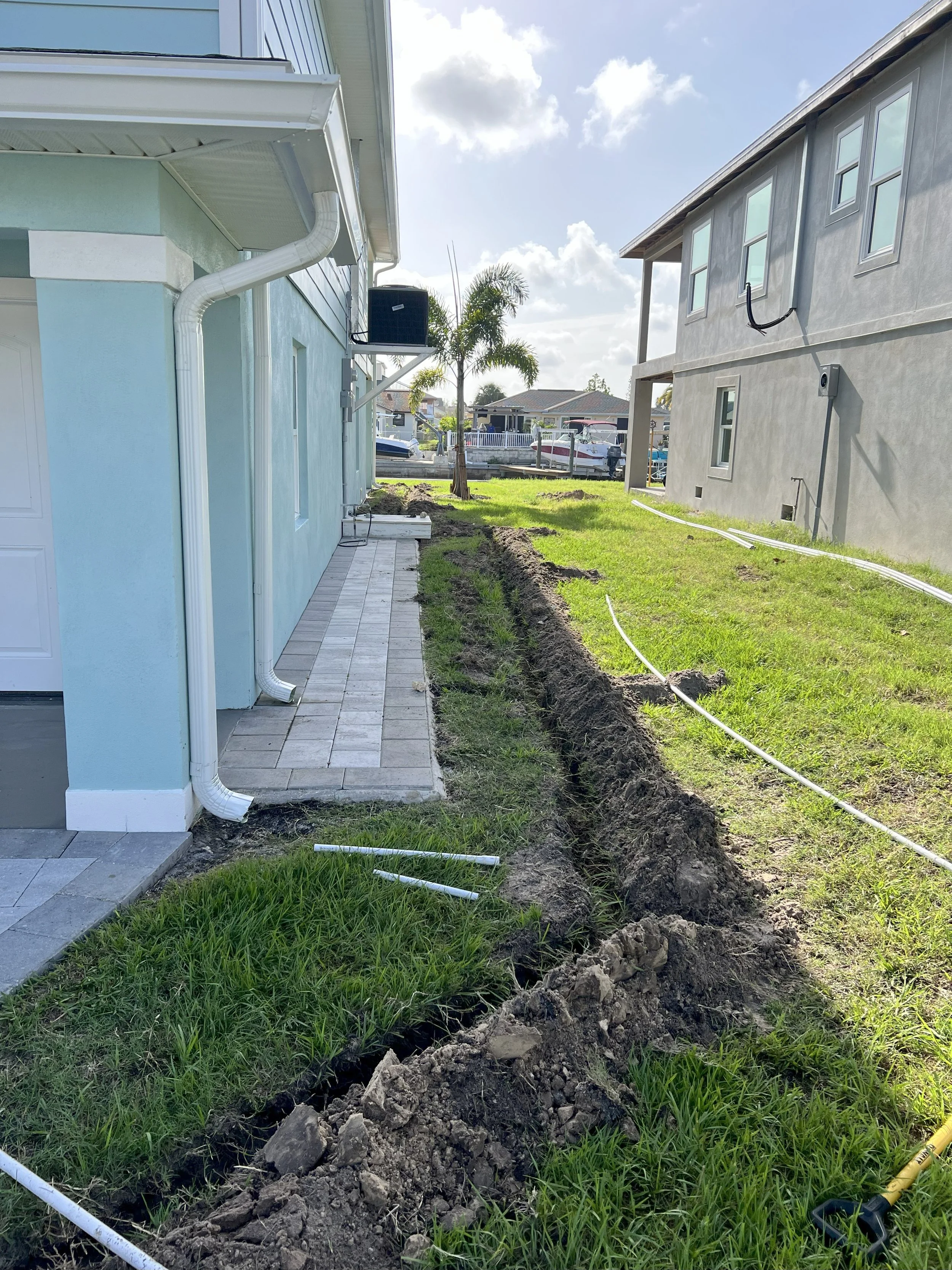 Excavation work along the side of a house with a trench dug into the ground, pipes laid in the trench, and a grassy yard nearby under a partly cloudy sky.