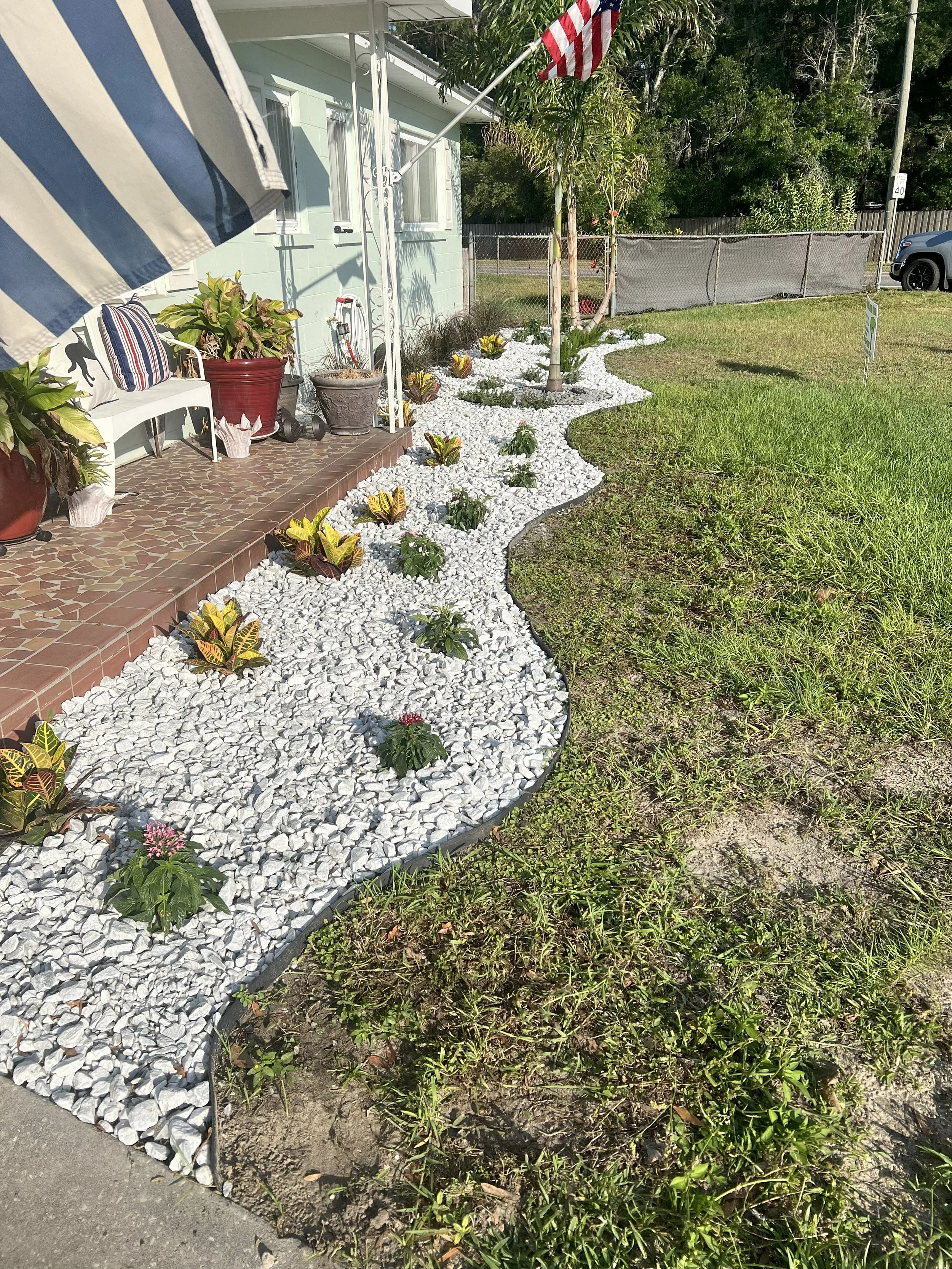 A front yard garden with a curved white gravel bed containing yellow and green leafy plants, surrounding a small tree. The garden is adjacent to a tiled porch with potted plants, a bench with striped pillows, and an American flag. The background has