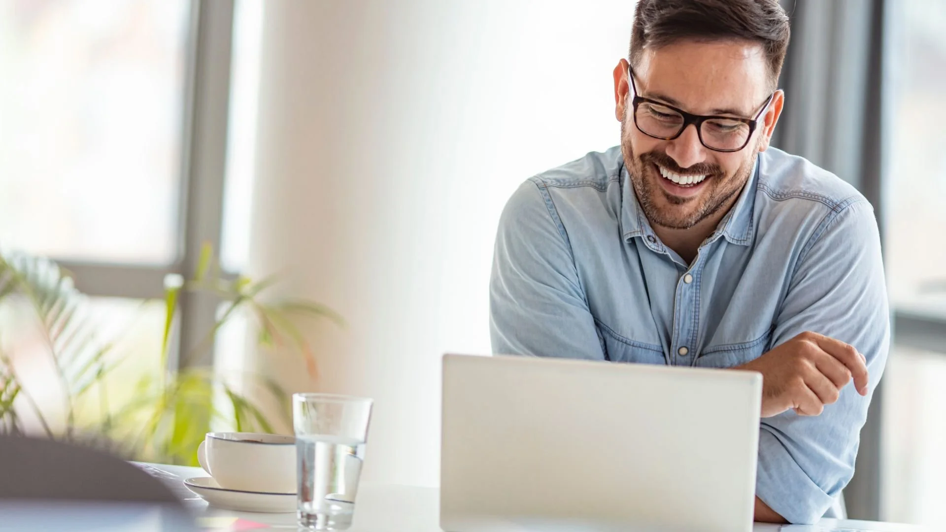 A woman wearing a brown turtleneck sweater is sitting at a table, smiling and looking at her laptop. She has earphones in and her hand is on her chest.