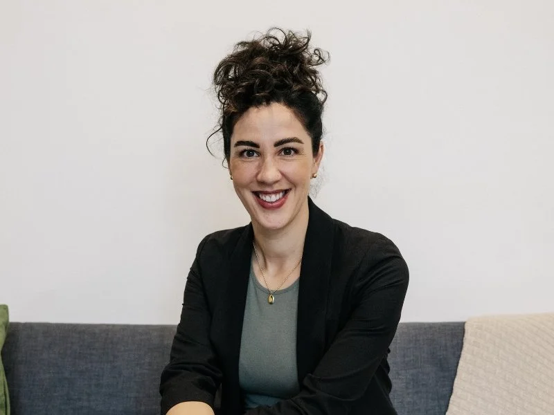 A woman with curly brown hair smiling, sitting on a gray couch with a white wall background.