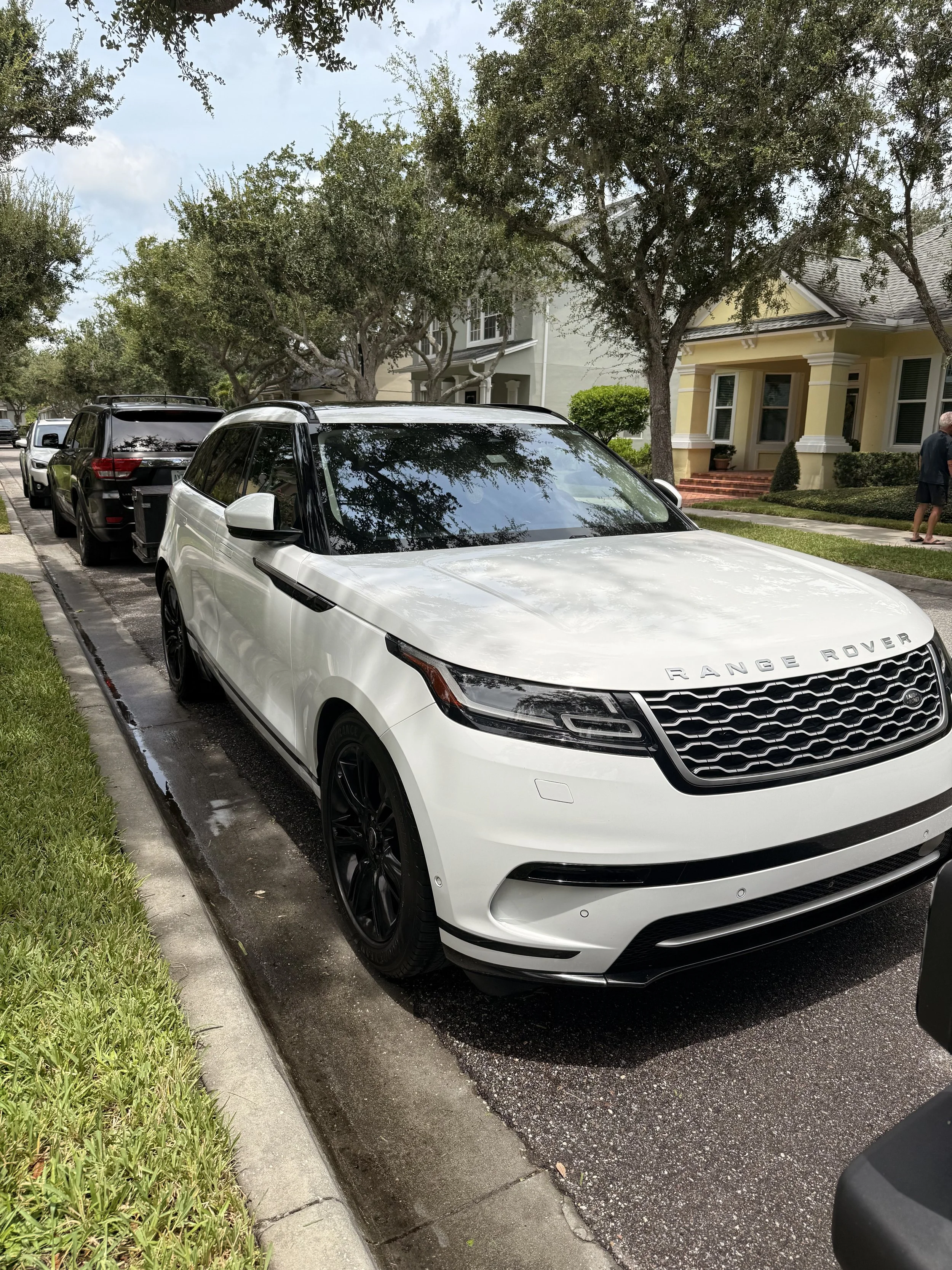 A white Range Rover parked on a residential street with other cars, houses, and trees visible in the background.