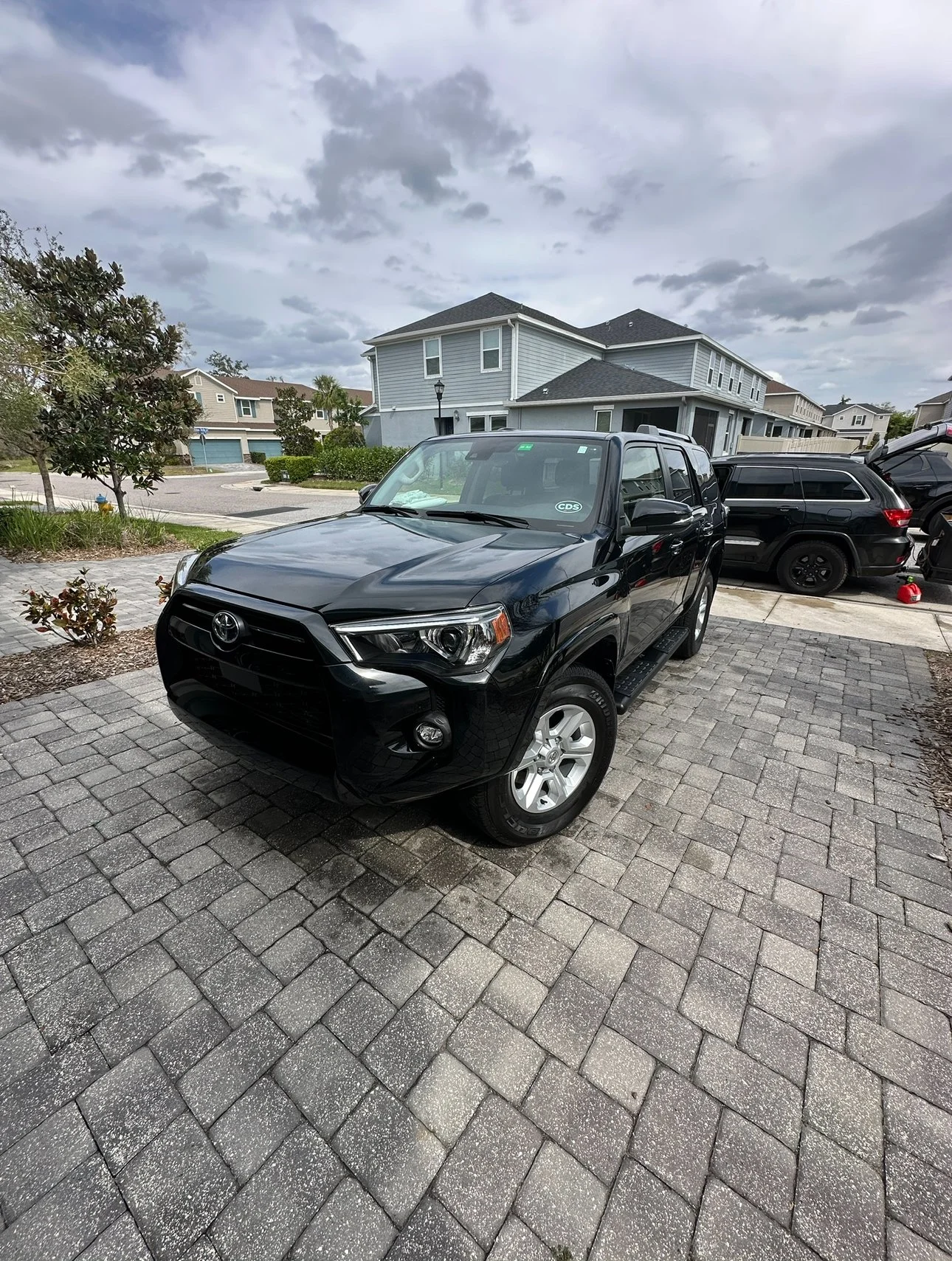 A black Toyota SUV parked on a brick driveway in a suburban neighborhood with residential houses and cloudy sky in the background.