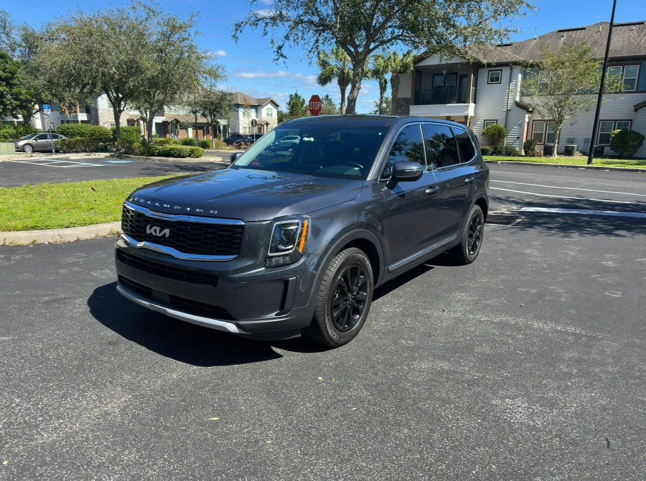 A dark gray Kia SUV parked in a parking lot with apartment buildings and trees in the background on a sunny day.