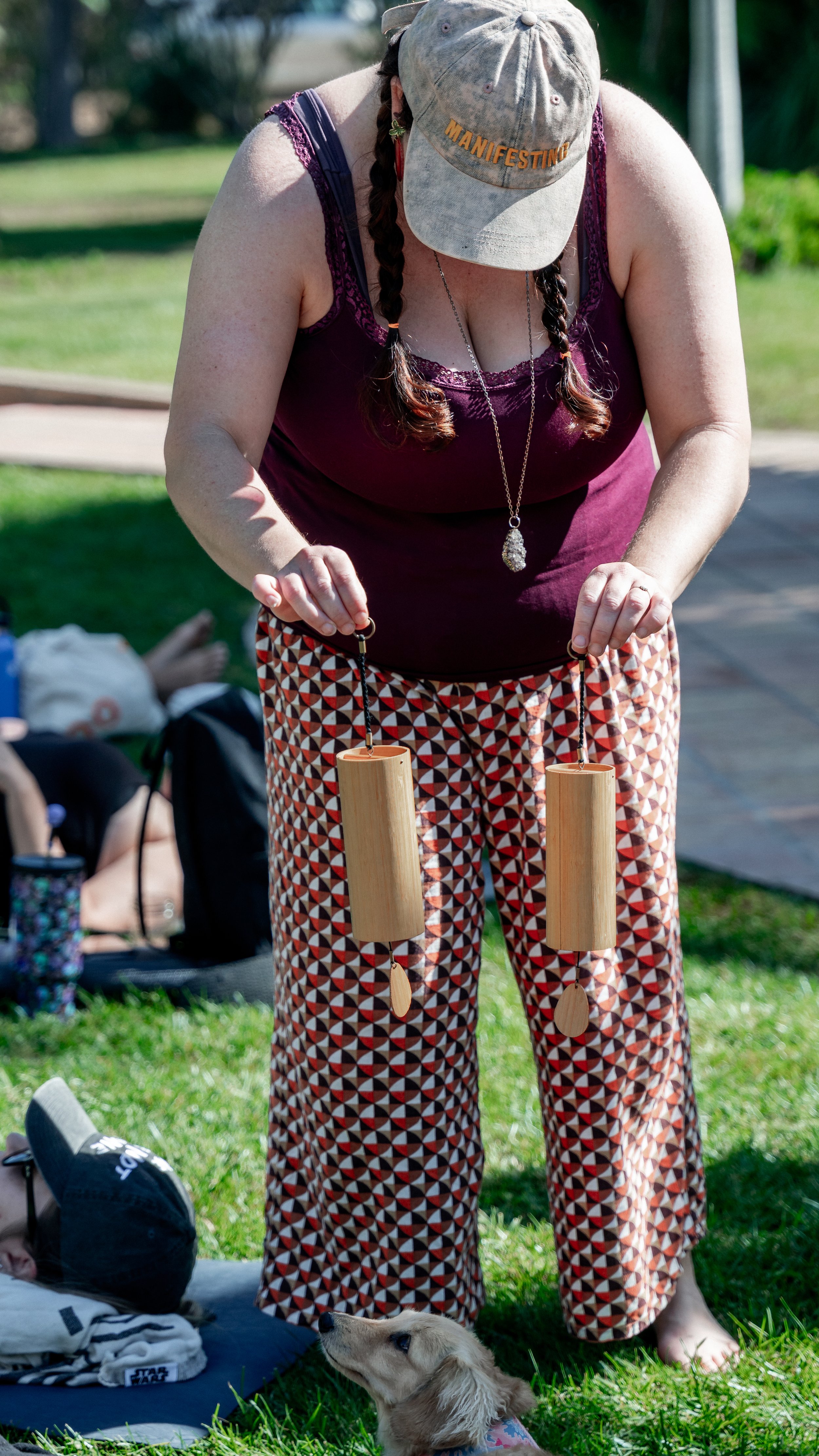 A woman with braided hair wearing a cap, purple tank top, and patterned pants, holding two wooden wind chimes outdoors on grass.