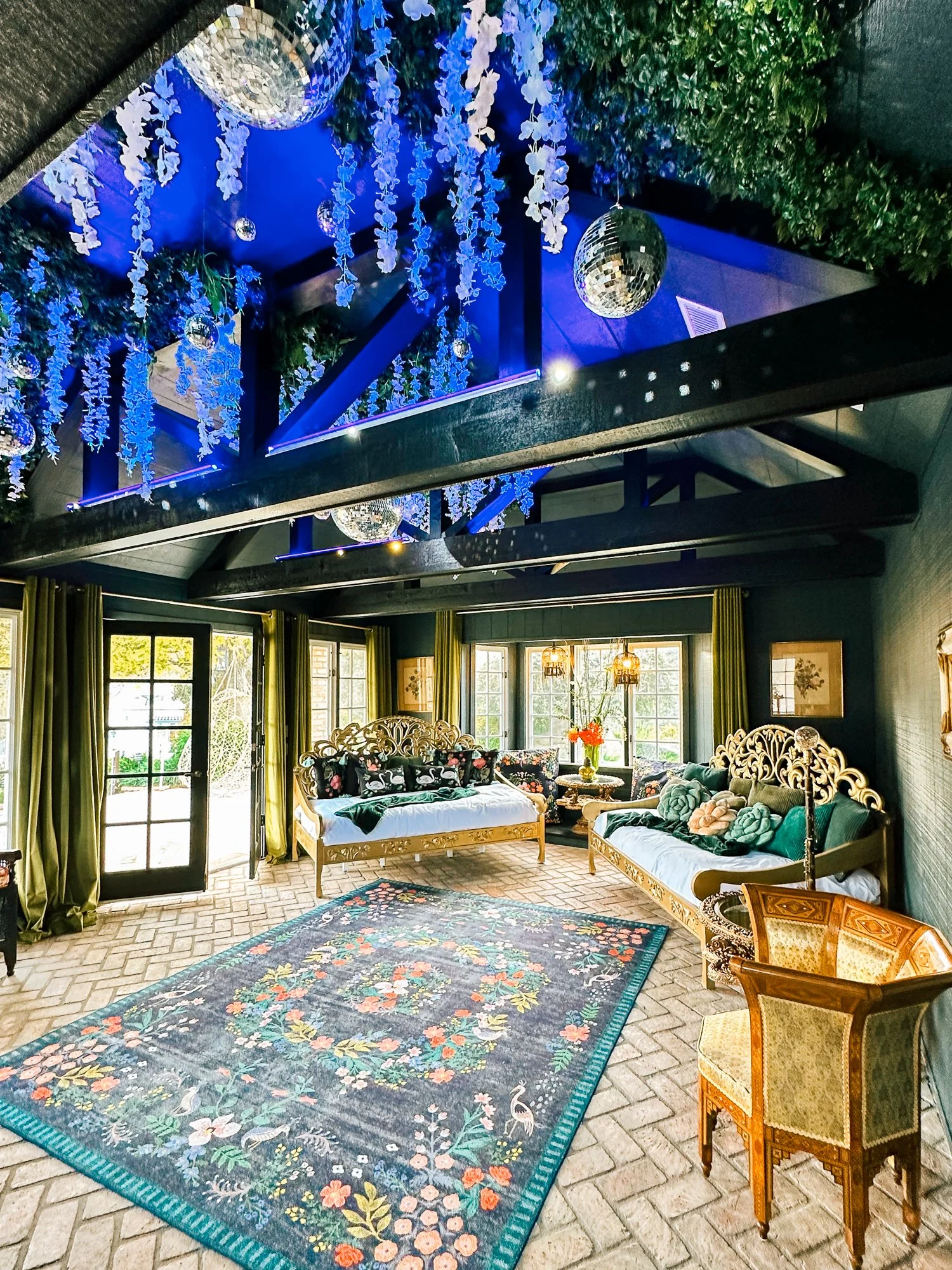 The image shows a cozy indoor living space with large windows and green curtains. There are two ornate wooden sofas with floral and animal-themed cushions, a smaller patterned wooden chair, and a colorful floral rug on a brick floor. The ceiling has a blue accent with hanging white and blue decorative elements and disco balls. The space is well-lit with natural light from the windows.