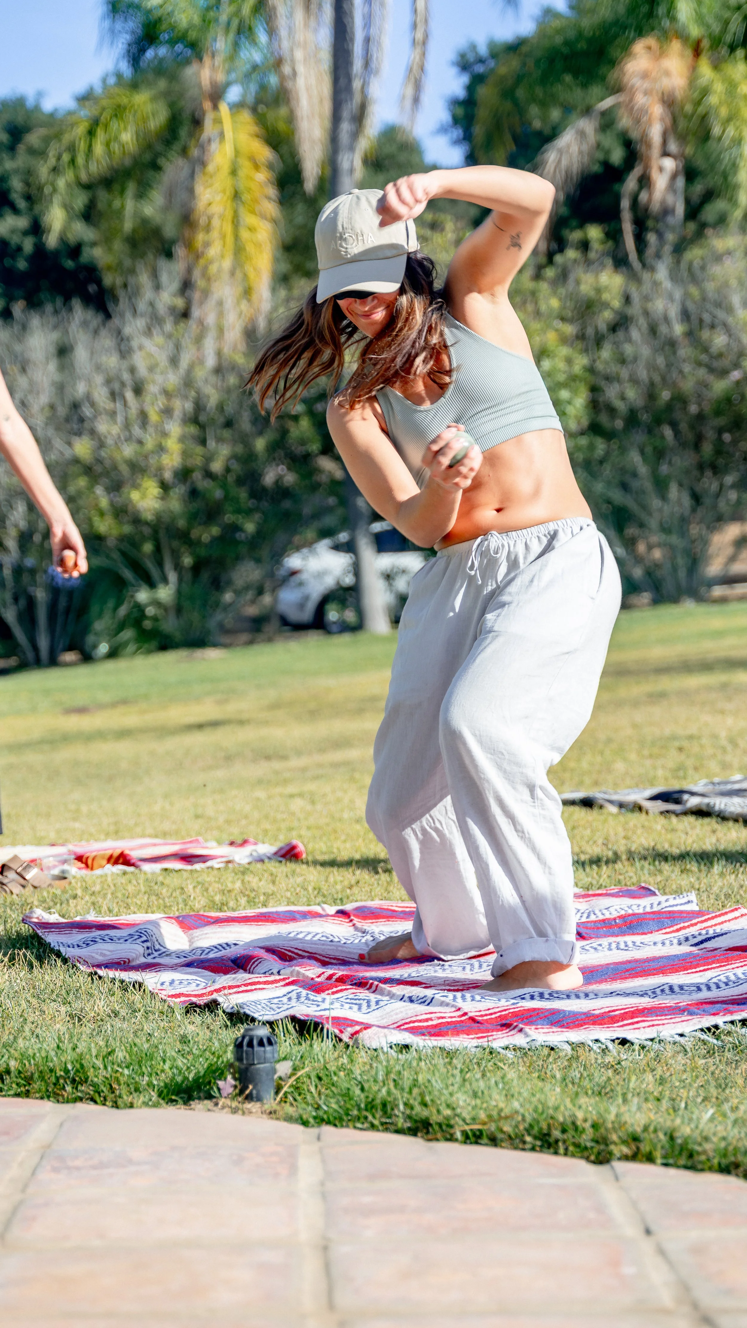 A woman with long brown hair wearing a gray crop top, white pants, and a beige cap is doing a yoga pose outdoors on a colorful striped blanket. There are trees and a blue sky in the background, and another person is partially visible on the left side of the image.