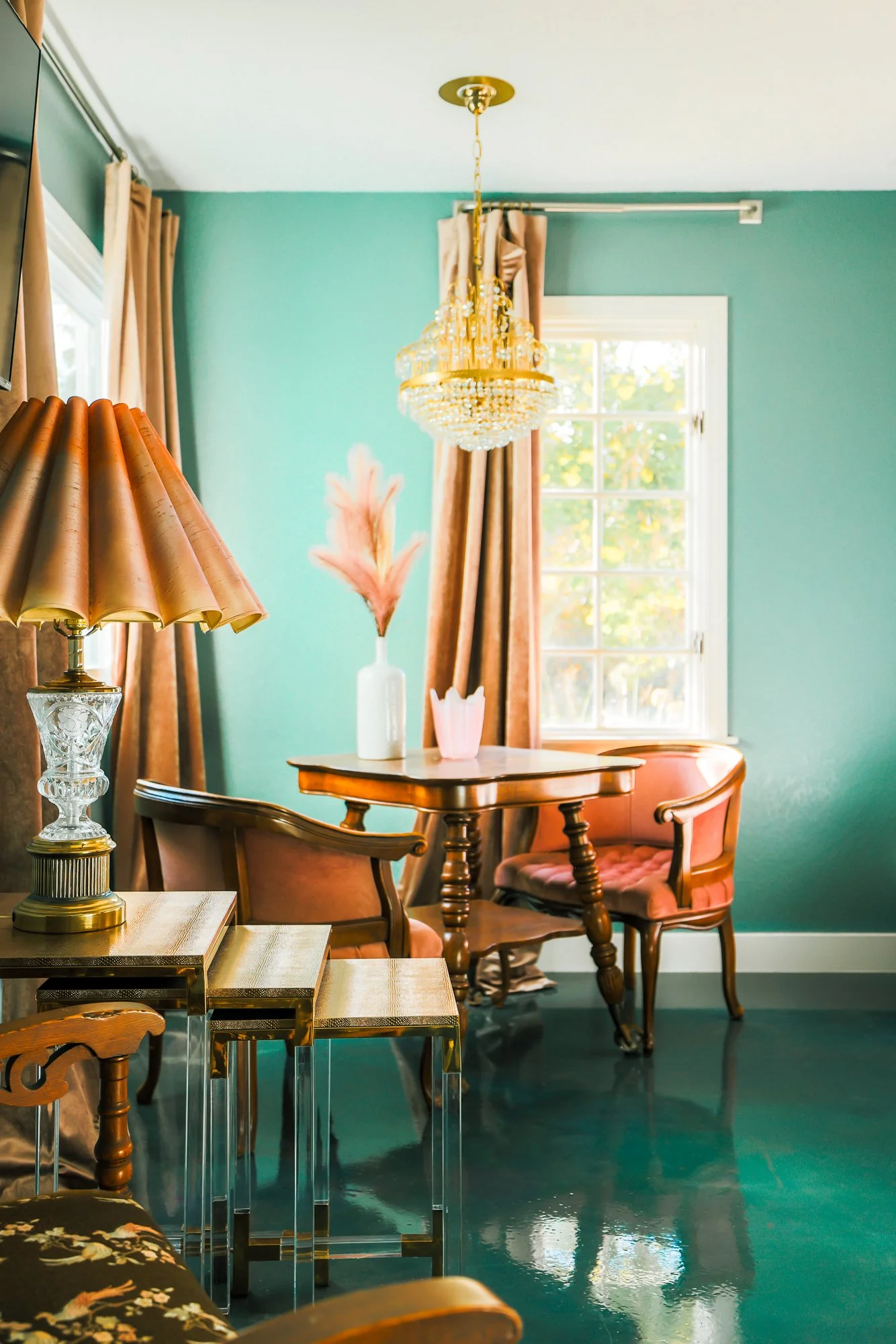 Elegant dining room with a wooden table and matching chairs, pink velvet upholstery, a vintage table lamp with a beige pleated lampshade, a pink decorative vase, a white vase with pink pampas grass, and a large window with beige curtains. A crystal chandelier hangs from the ceiling, illuminating the room with natural light from the window. The walls are painted teal, and the floor is shiny dark hardwood.