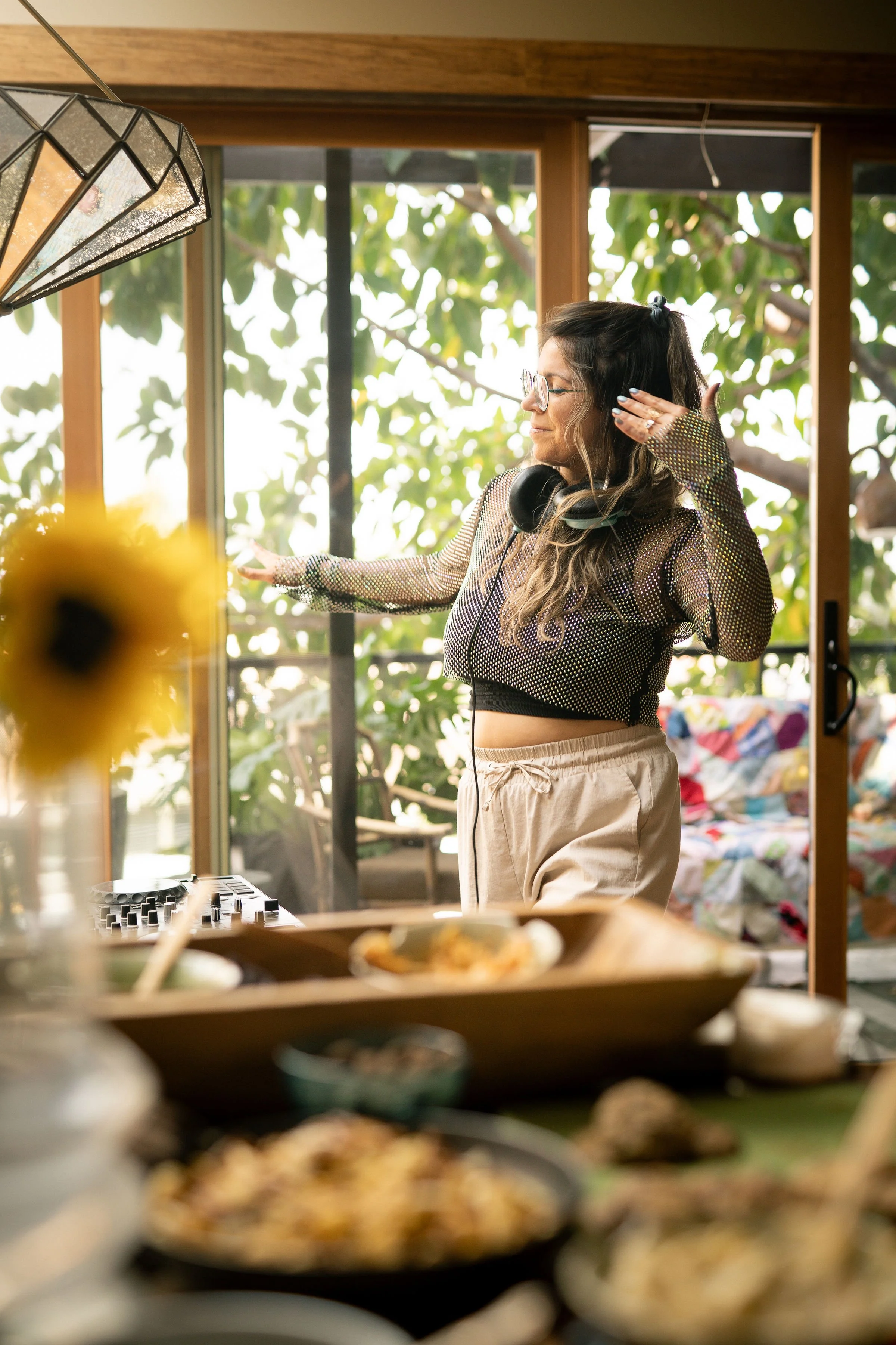 A woman with glasses and wavy hair wearing a mesh top and beige pants, wearing headphones, dancing while DJing in a bright room with large windows and green outdoor scenery.