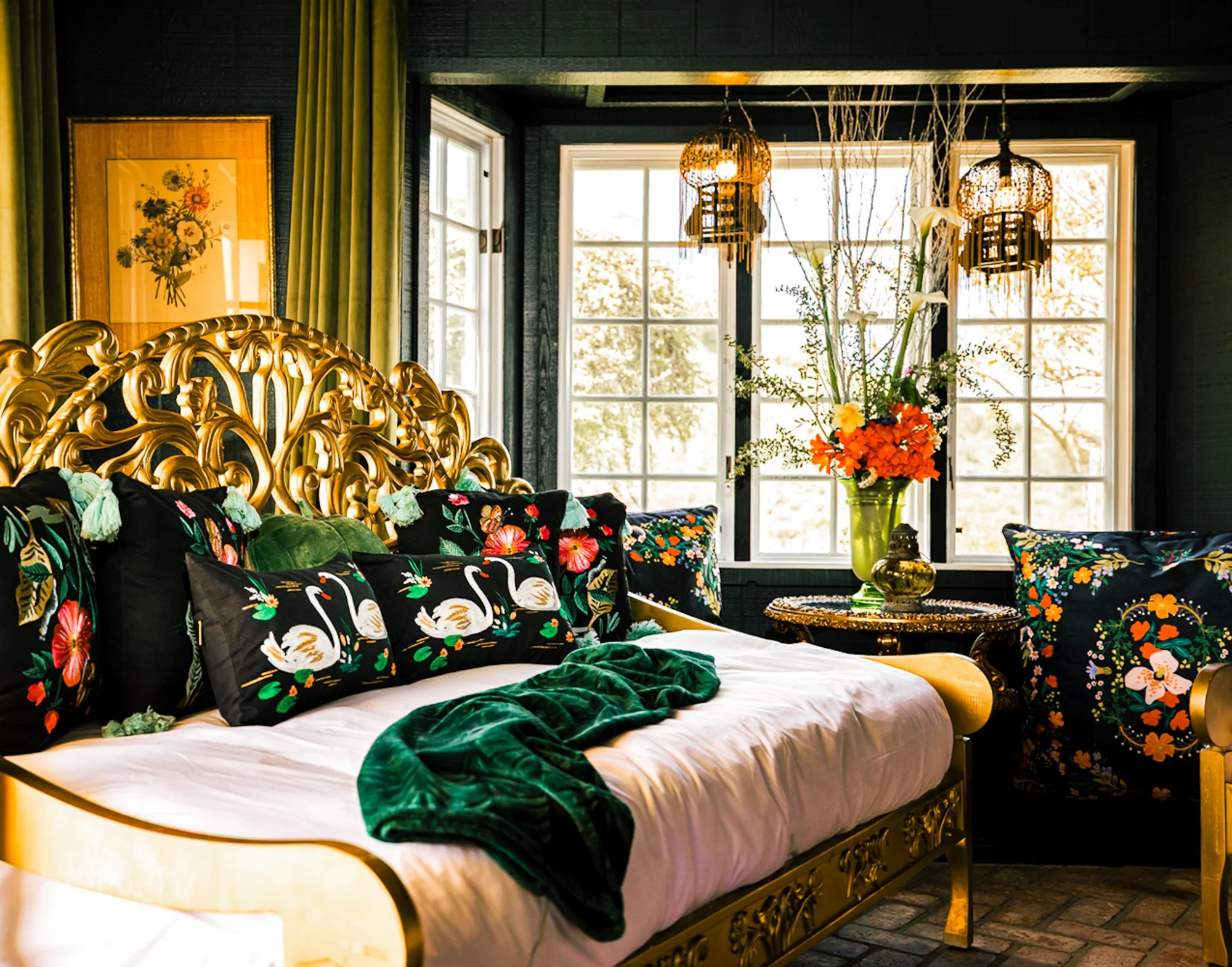 Luxurious bedroom with a gold ornate bed frame, black floral bedding, and swan pillows. Large window with green curtains, framed floral artwork, hanging wicker lamps, and a glass vase with white lilies on a side table.