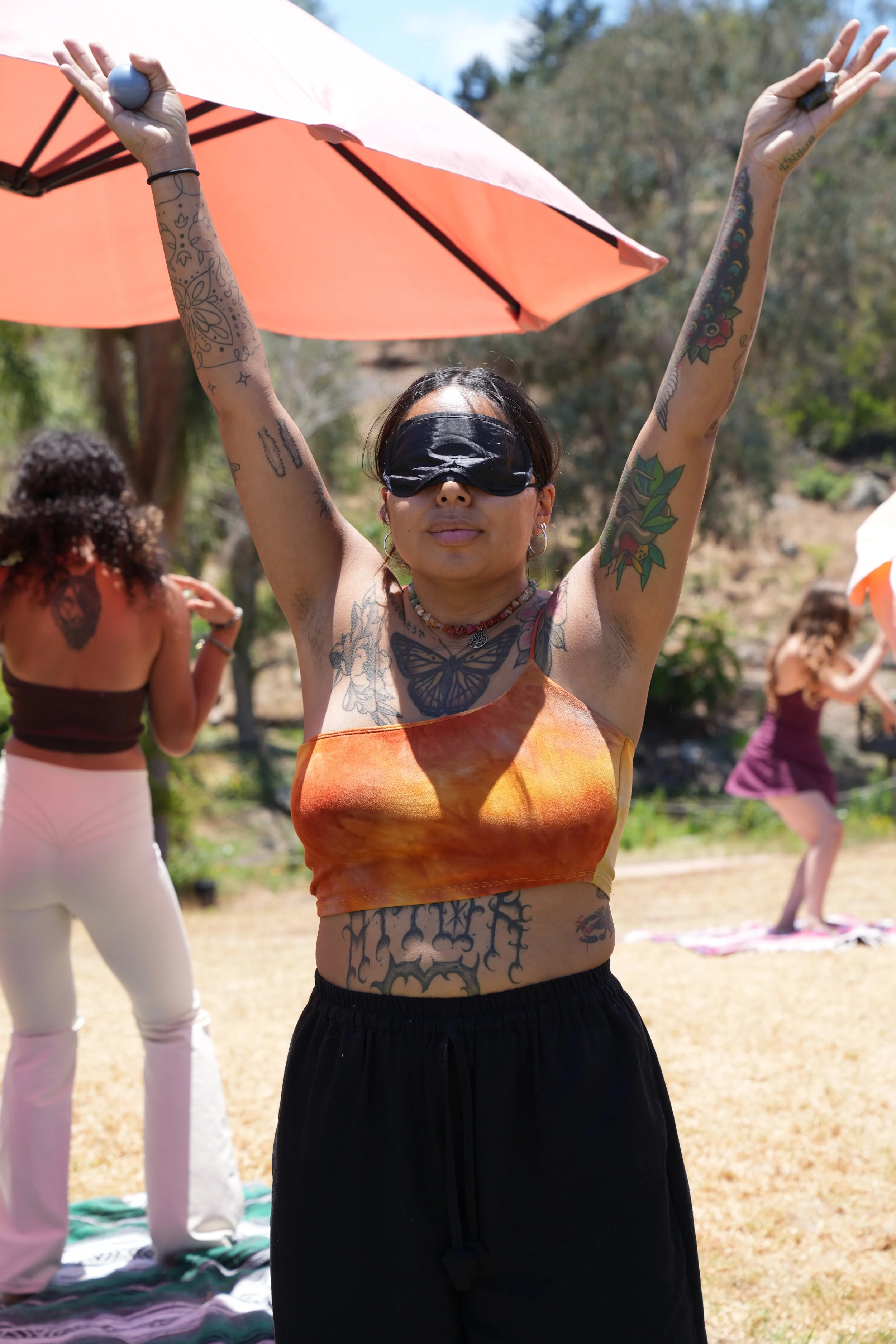 A woman with tattoos practicing yoga outside in a park, wearing a black blindfold, orange tie-dye crop top, and black pants, with her arms raised holding small objects, under a pink and orange umbrella.