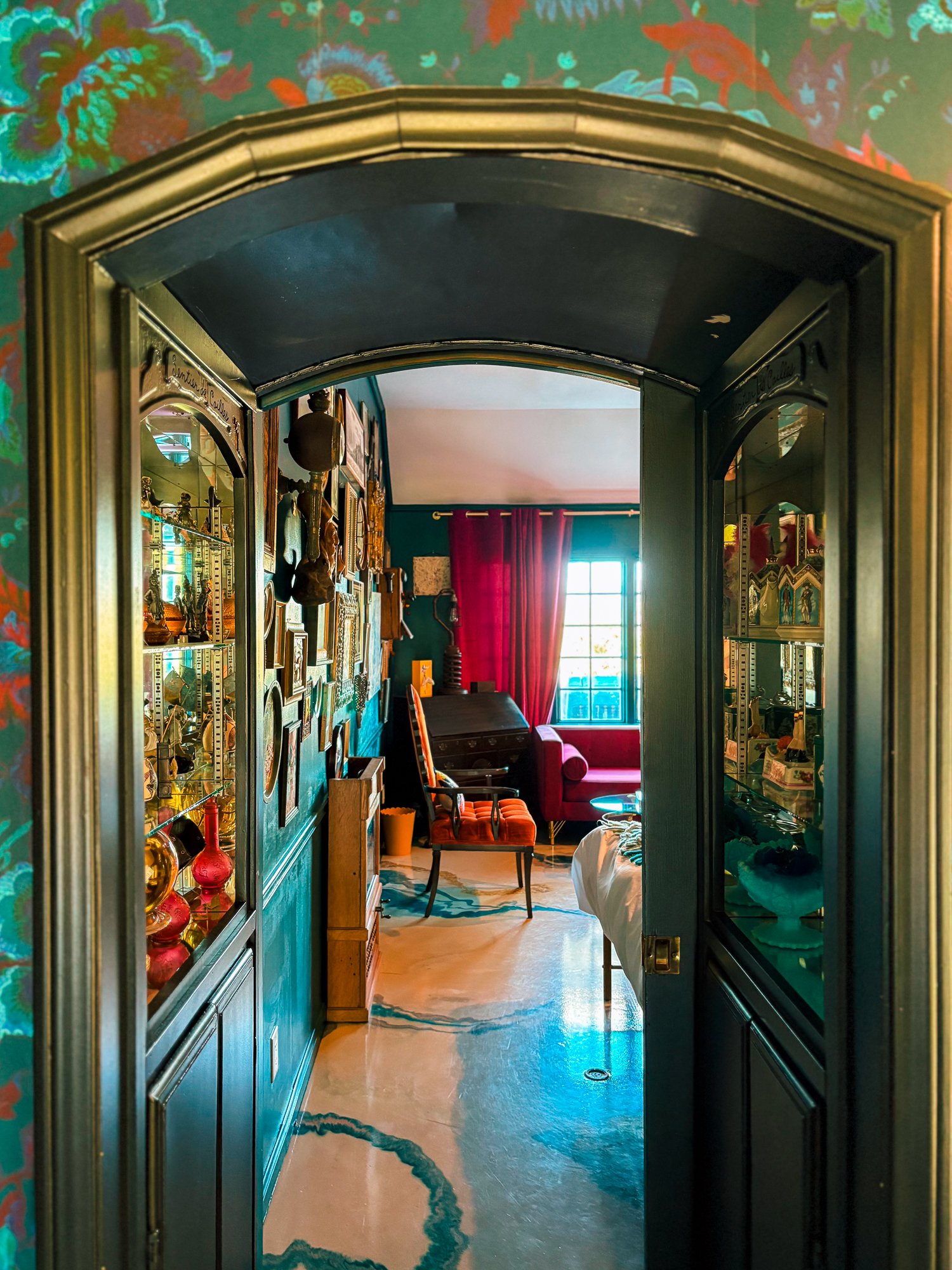 View through a doorway into a cozy room filled with eclectic decor. The room features a window with red curtains, a red sofa, and a wooden chair with an orange cushion. The wall has various artwork and decorative objects. The floor is marble with a greenish-blue vein pattern, and a large mirror reflects part of the room. The doorway frame is ornate with a gold finish.