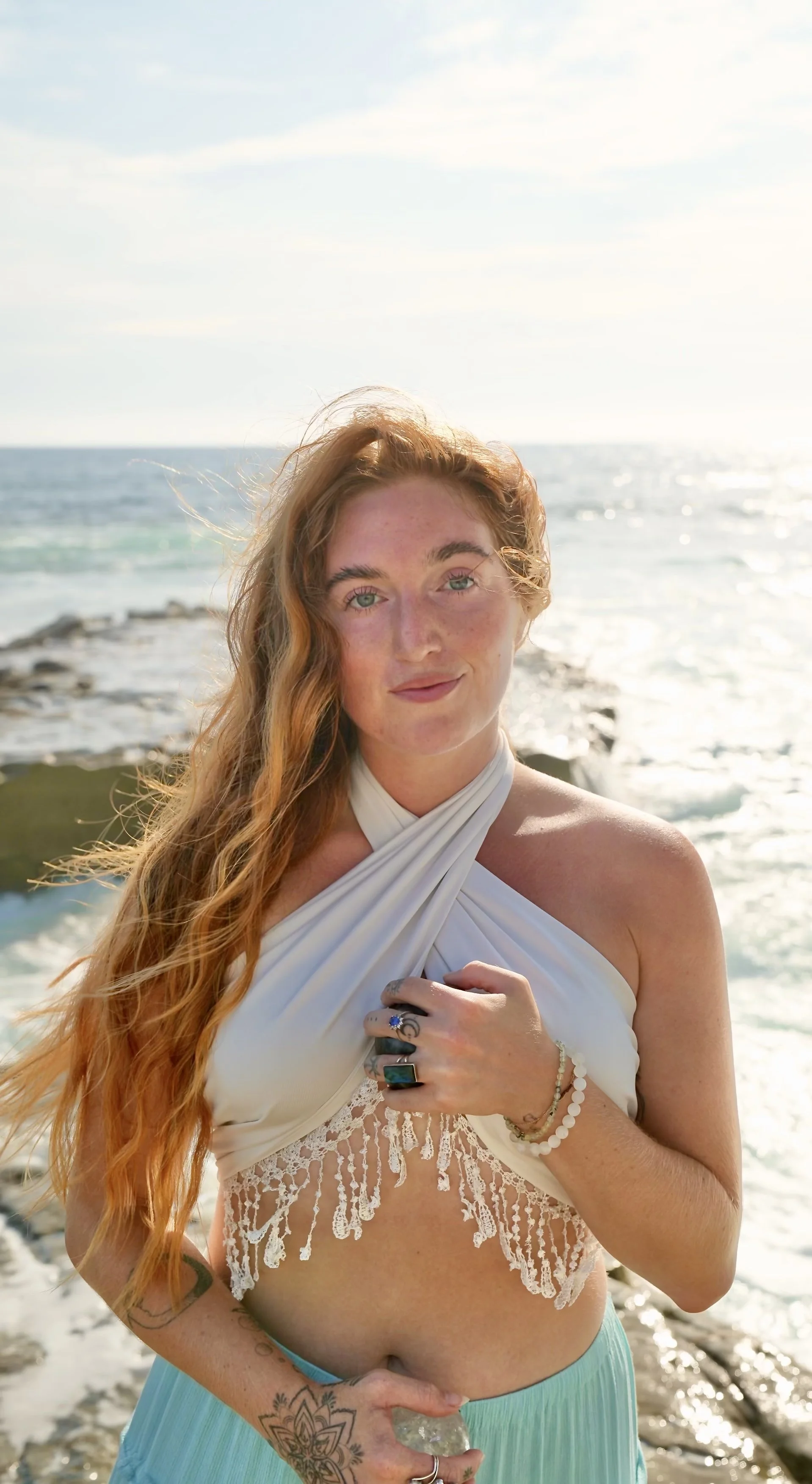 A woman with long, wavy red hair and green eyes stands on a beach near the ocean, wearing a white halter top with lace detail and a light blue skirt. She has multiple rings, bracelets, and tattoos, and is holding a clear quartz crystal. The background shows water, rocks, and a partly cloudy sky, with sunlight reflecting off the ocean.