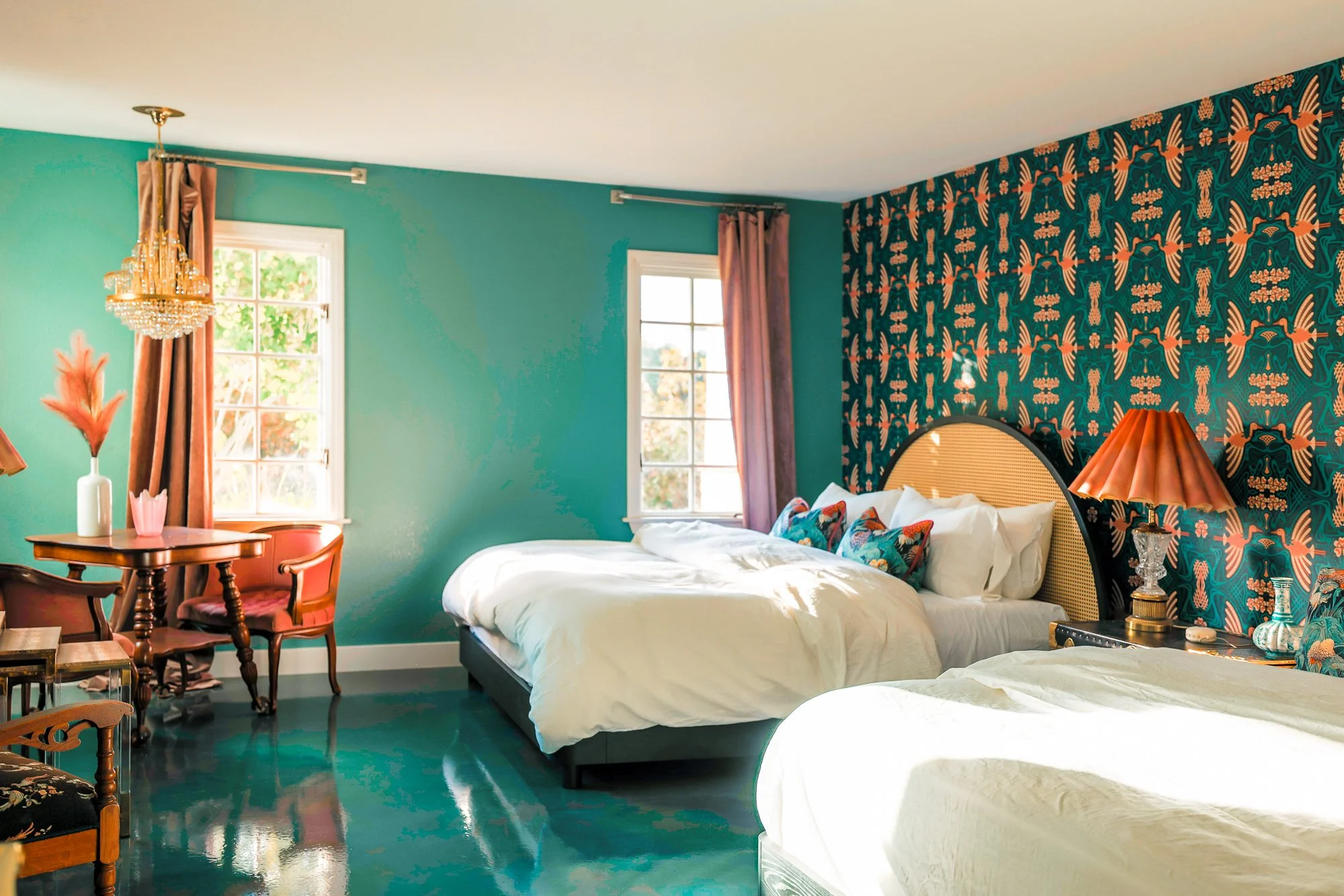 A colorful bedroom features a teal wall with patterned wallpaper, two windows with pink curtains, a chandelier, a bed with white bedding and patterned pillows, a wooden table with a vase of pink feathers, and a bedside table with a lamp.