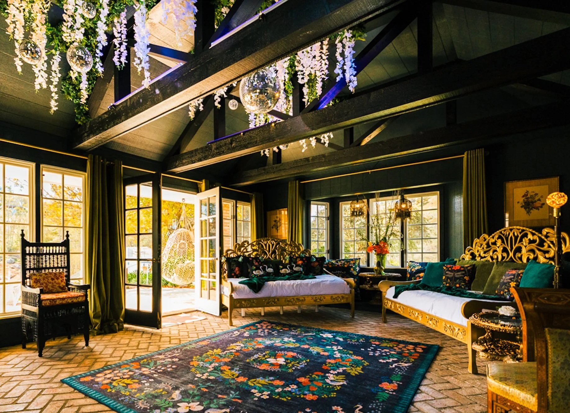 A bright living room with large windows, green curtains, and a high ceiling with black beams, decorated with hanging white flowers and mirrored disco balls, featuring ornate furniture, colorful cushions, a floral rug, and decorative lamps