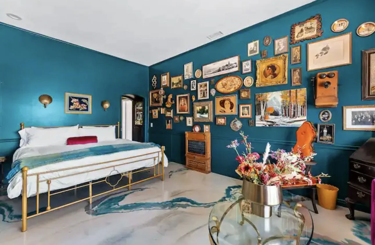 A bedroom with a teal wall decorated with various framed pictures, a gold bed with white bedding and a red pillow, a wooden chair with orange upholstery, a floral arrangement on a glass table, and a window reflecting trees.
