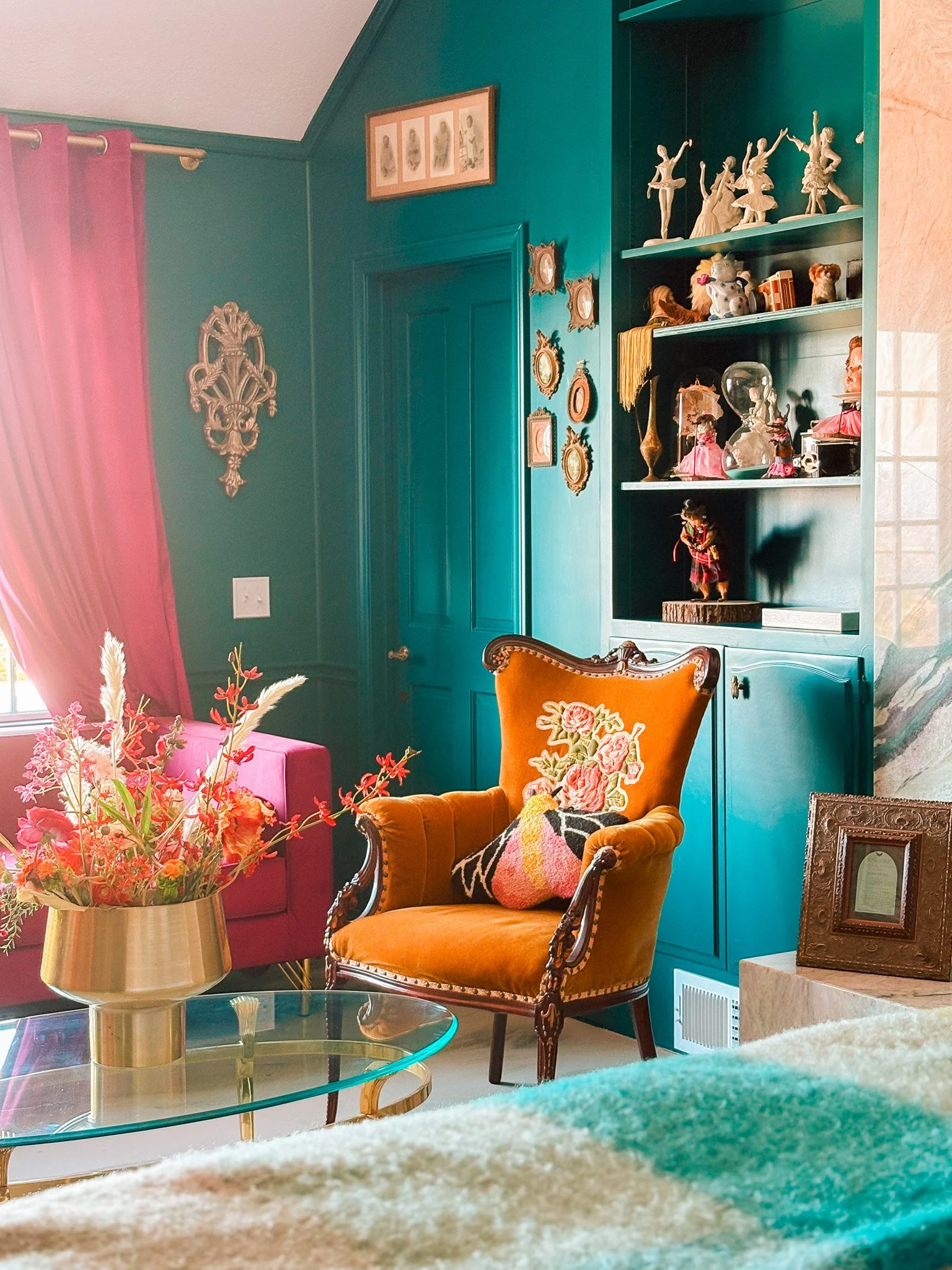 A cozy living room with a teal wall and a glass coffee table. A vintage orange armchair with embroidered pillows is in front of the table. A large pink candelabra with flowers sits on the table. The room has a built-in teal bookshelf decorated with figurines and collectibles. Pink curtains frame a window on the left, and various framed artworks and decorative mirrors are hung on the walls.