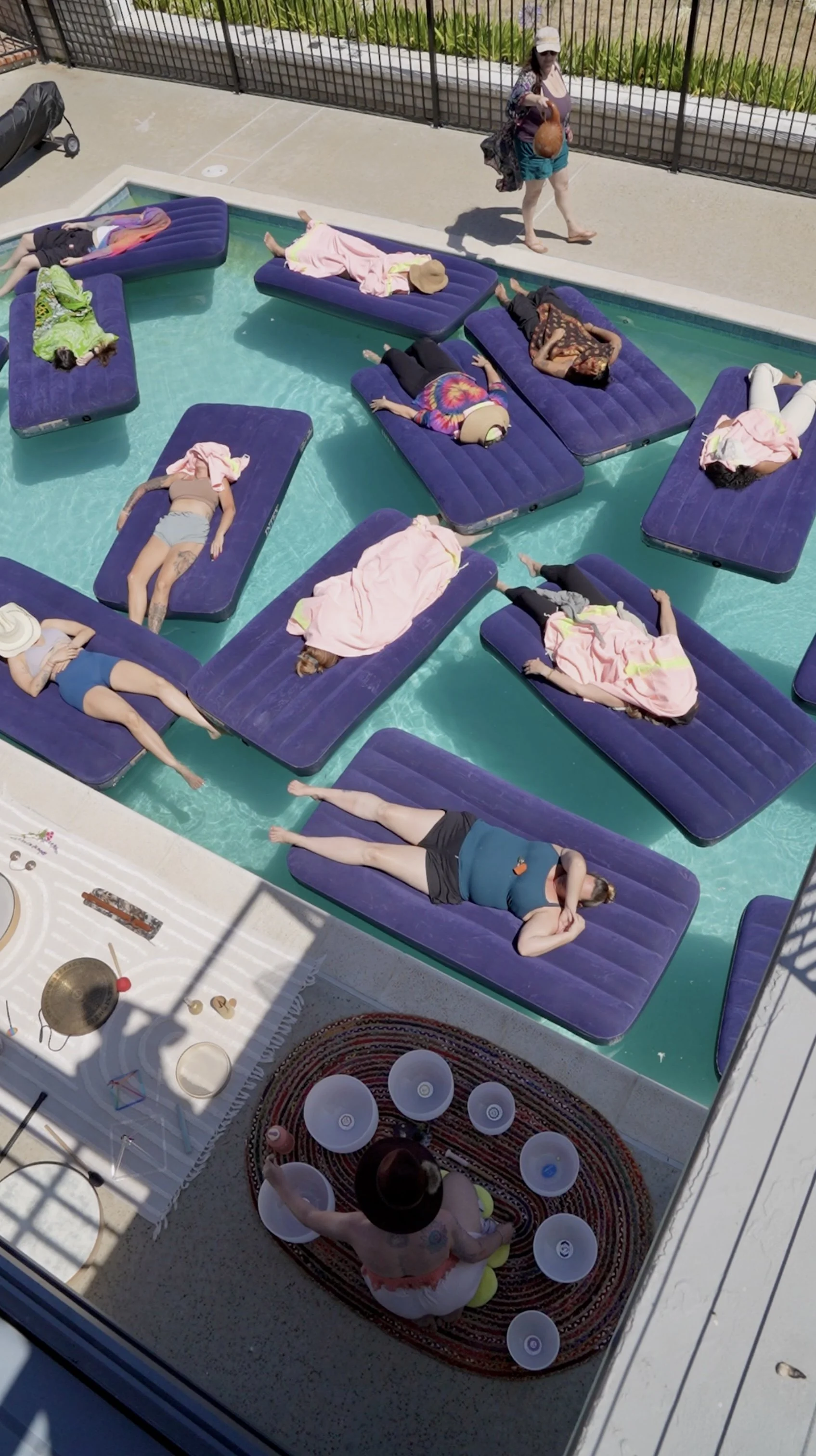 People are relaxing on pool float loungers inside a swimming pool, with some lying down and others sitting or standing nearby. There is a woman sitting on a striped round rug, playing with plastic bowls, surrounded by sunlit shadows.