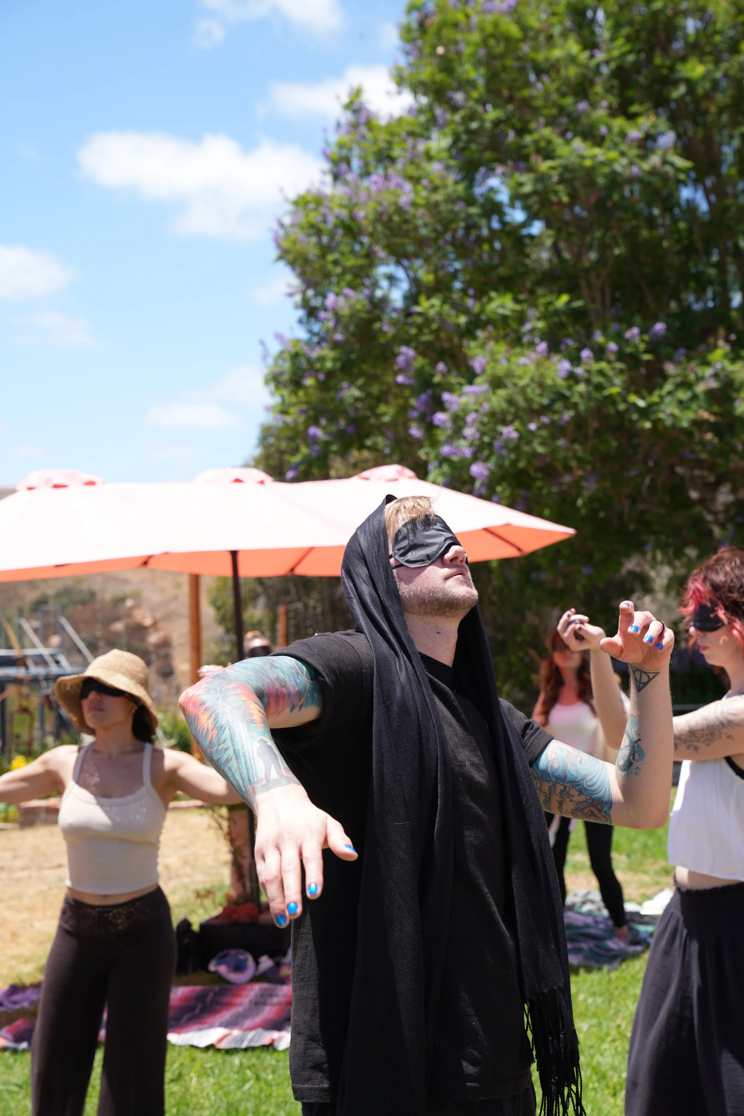A man with tattoos and a blindfold doing a yoga pose outdoors. Women with hats and tattoos following along, with a large tree and blue sky in the background.