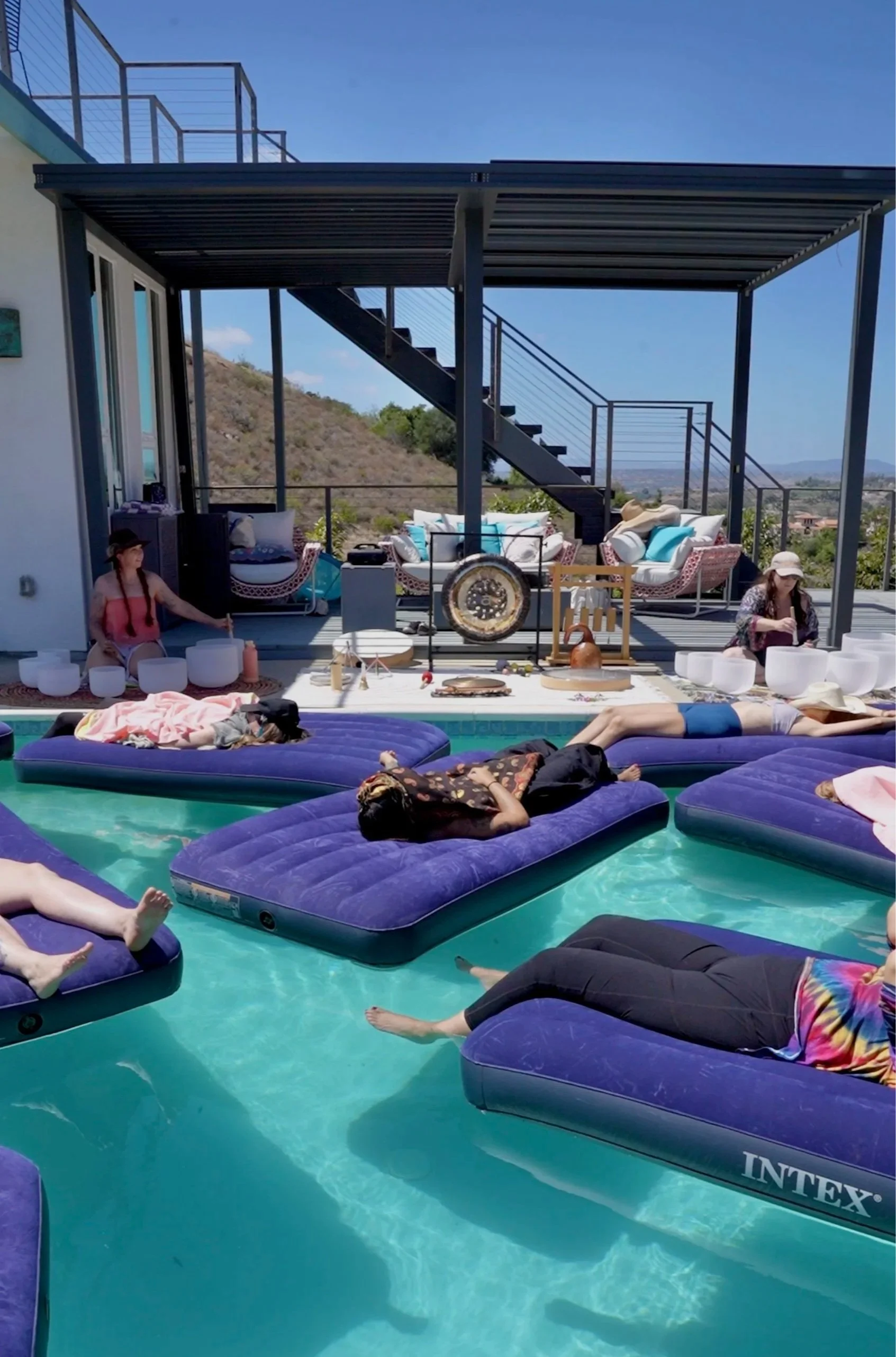 People relaxing on purple inflatable mattresses in a swimming pool, with some lying down and others sitting, under a clear blue sky.