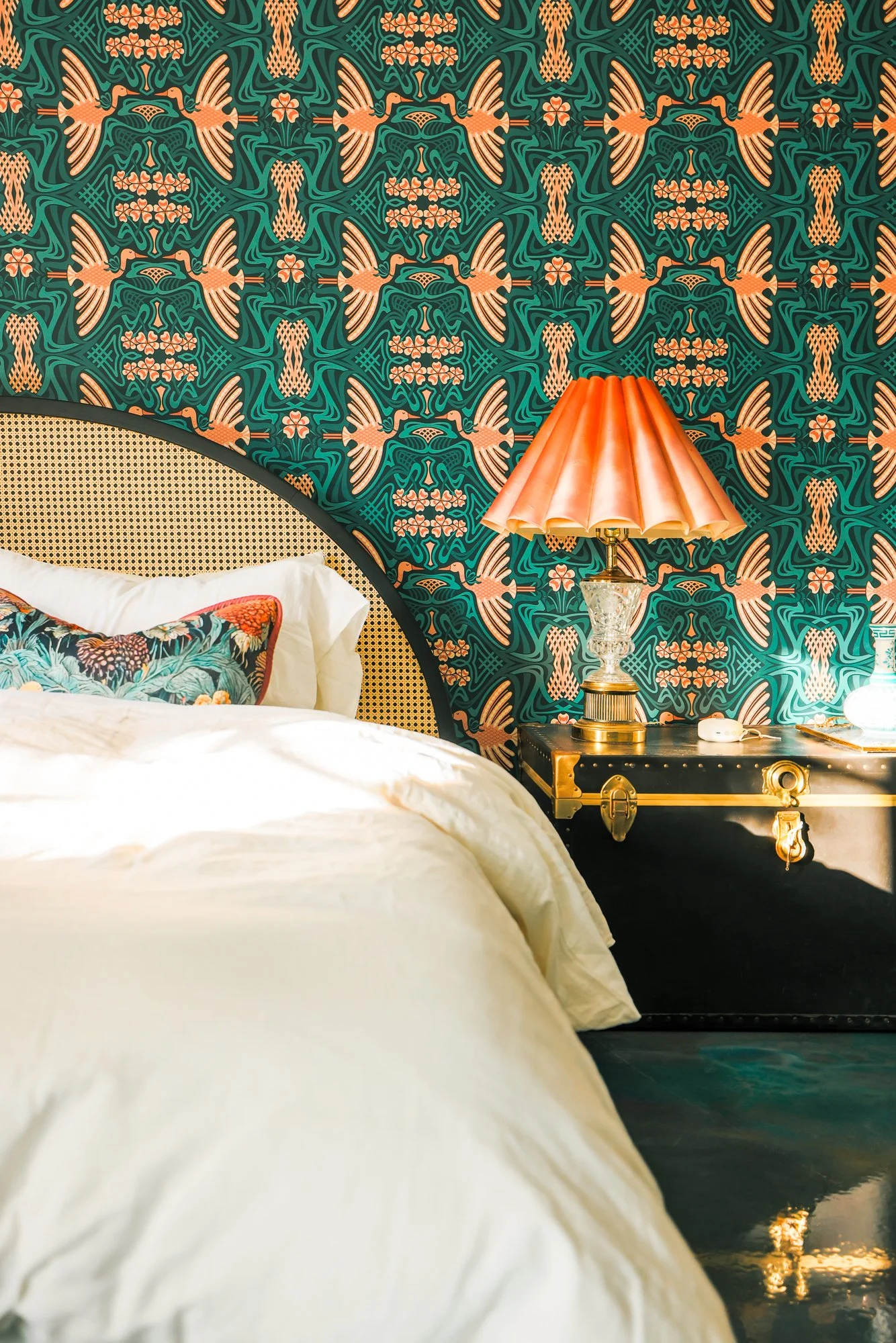 A bedroom scene featuring a bed with white linens and decorative pillows, a patterned wallpaper with birds and floral designs, a black nightstand with a gold-trimmed black surface, a ornate table lamp with a copper-colored shade, a glass base, and a black and gold decorative box on the nightstand.