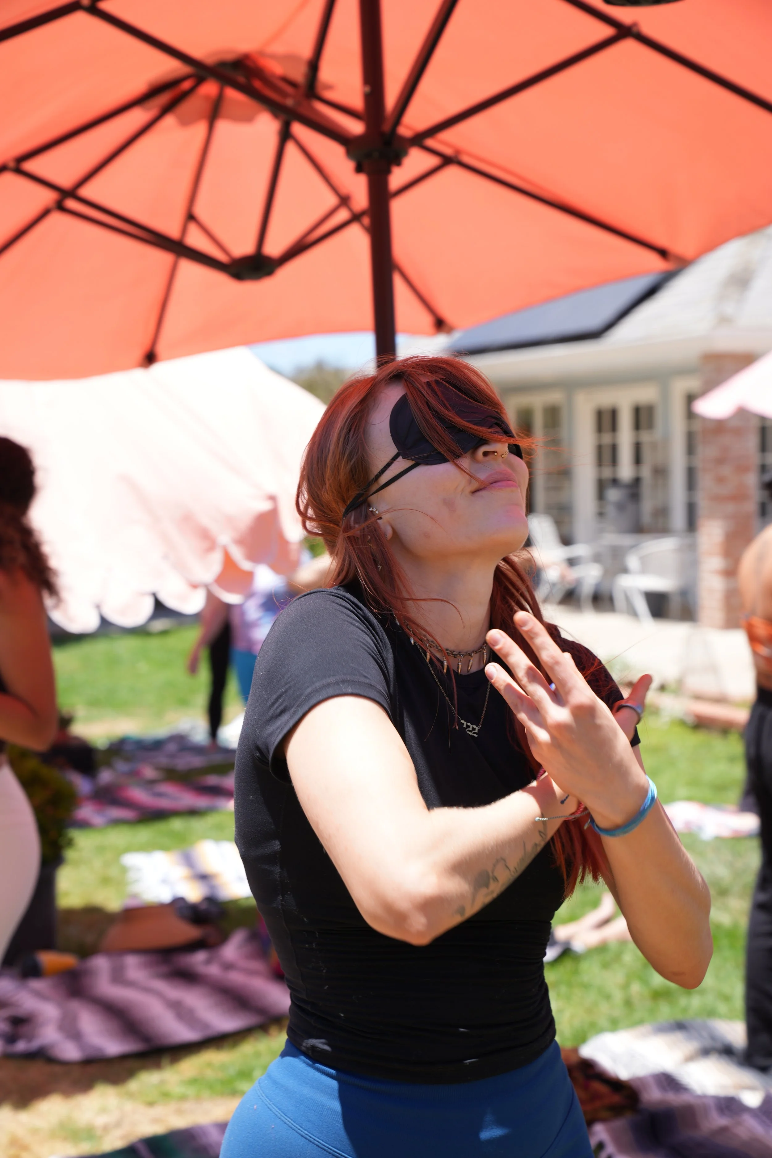 A woman with red hair wearing a black eye mask practices yoga outdoors under a large pink umbrella, with people and blankets on the grass in the background.