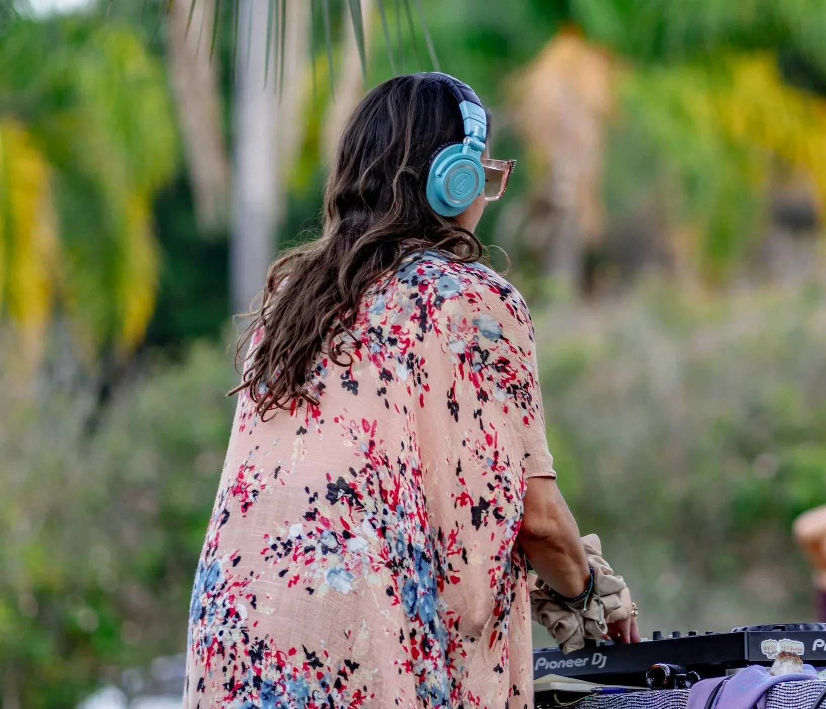 A woman with long wavy hair wearing glasses and blue headphones standing outdoors near trees, with a DJ mixer and a laptop on a table beside her.