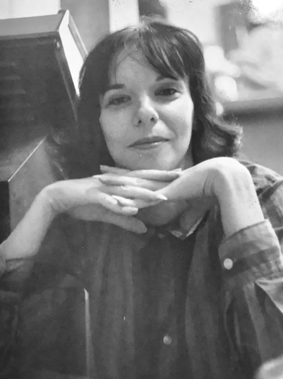 Black and white photo of a woman with dark, shoulder-length hair, smiling slightly, resting her chin on her folded hands on a table, sitting in a casual setting.