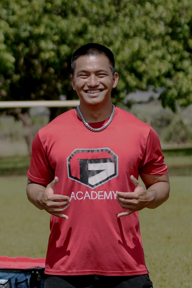 A young man wearing a red T-shirt with a logo and 'ACADEMY' written on it, smiling and making a hand gesture, standing outdoors on a grassy field with trees in the background.