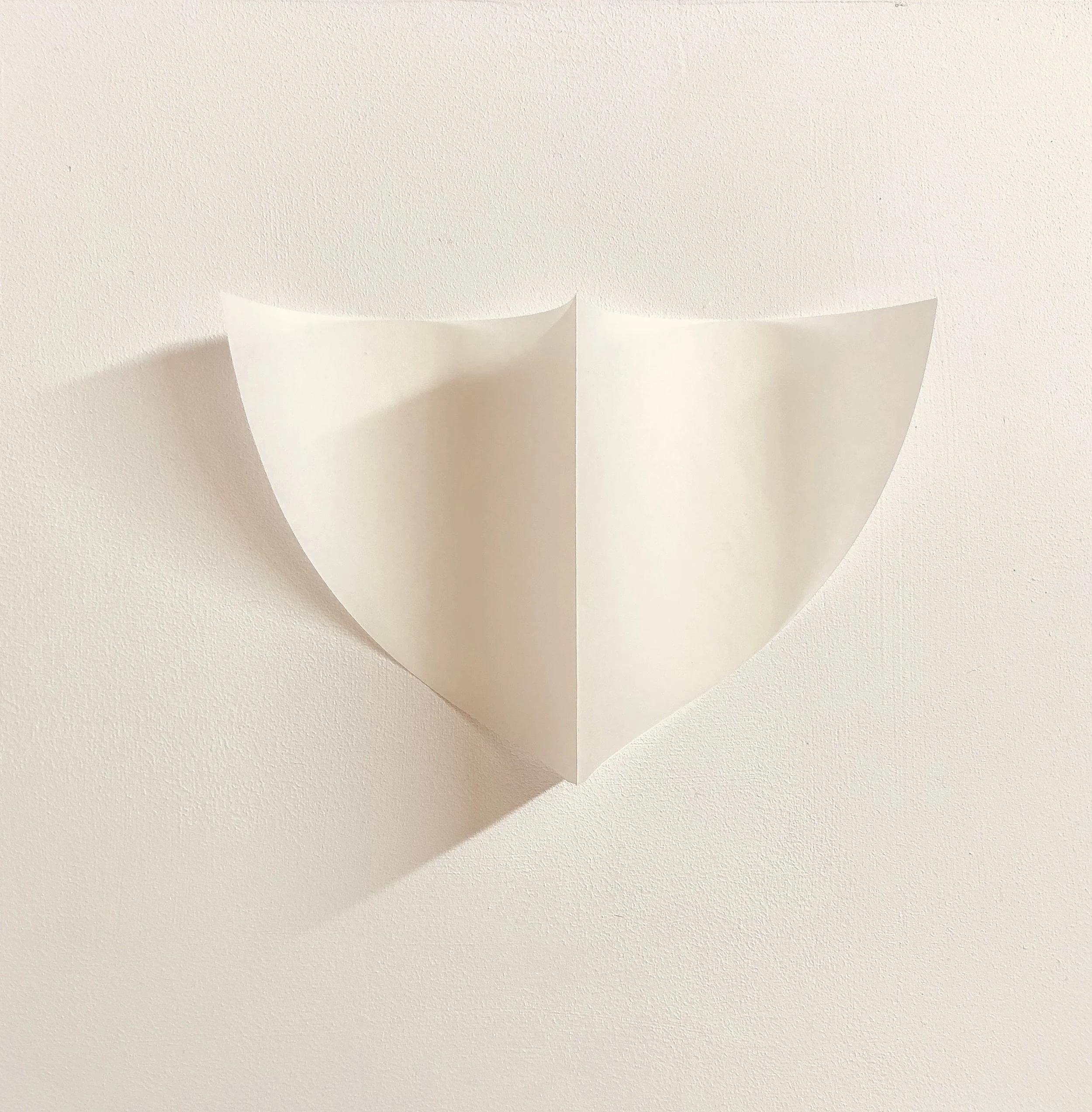 A white paper folded in a shield shape on a white wall, with shadow cast on the wall.