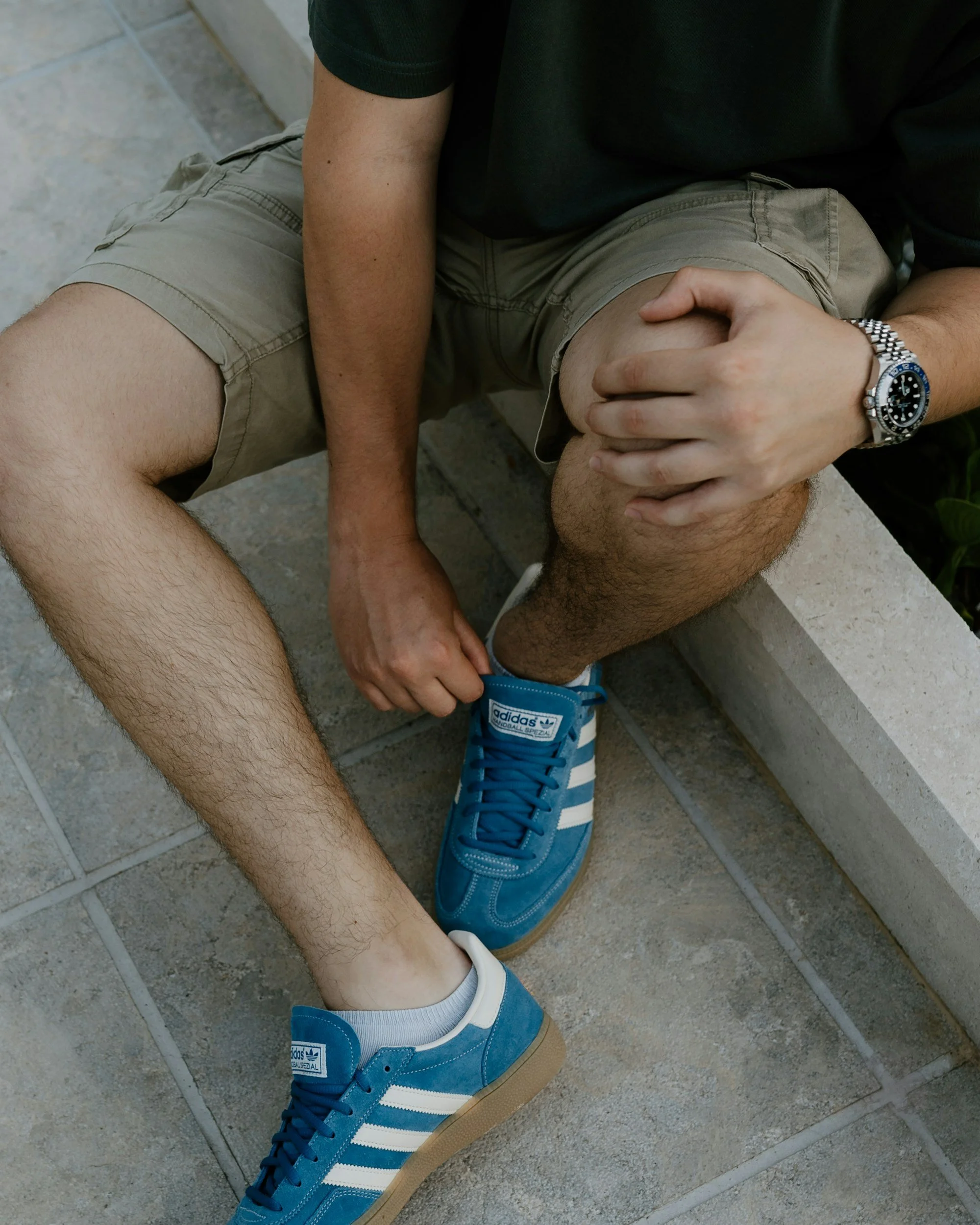 A man sitting on a bench tying the shoelaces of his blue Adidas sneakers, wearing khaki shorts and a black T-shirt.