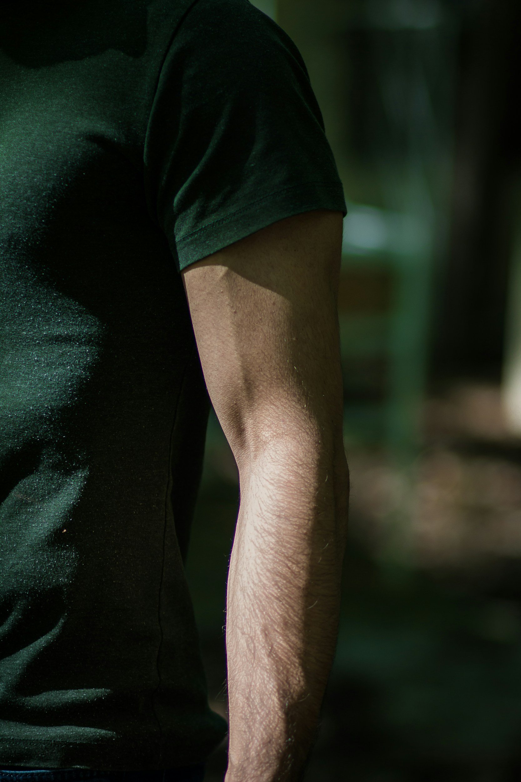 A person wearing a dark green shirt, with focus on their chest, shoulder, and arm, in an outdoor setting with blurred background.